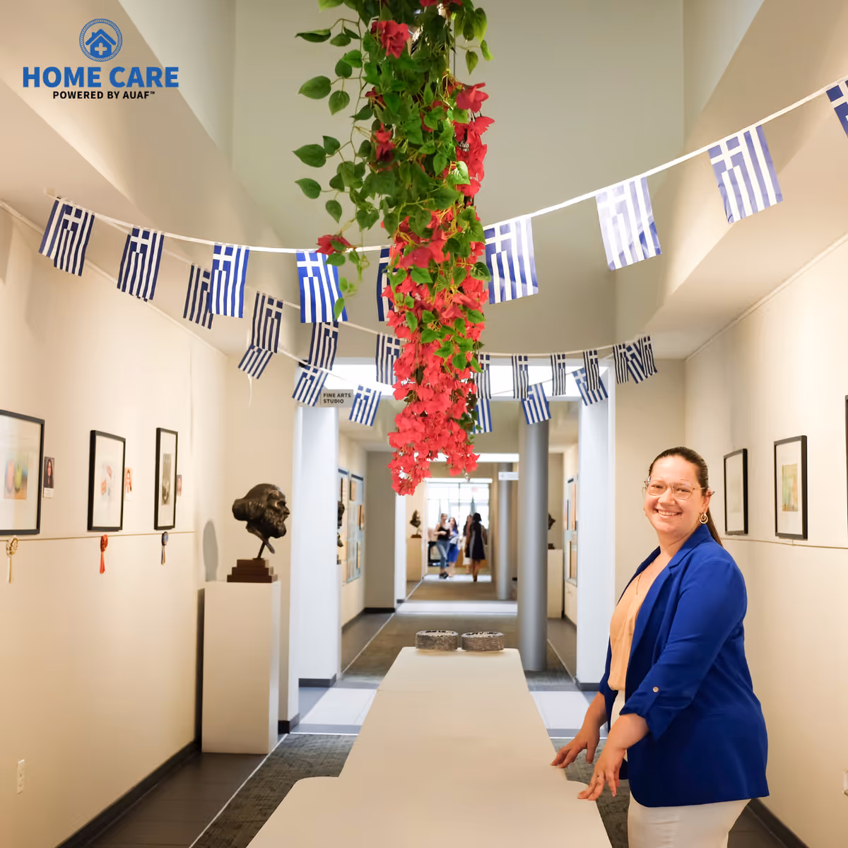 Bright facility hallway with a long table, Greek flags, hanging plants, and a smiling staff member.