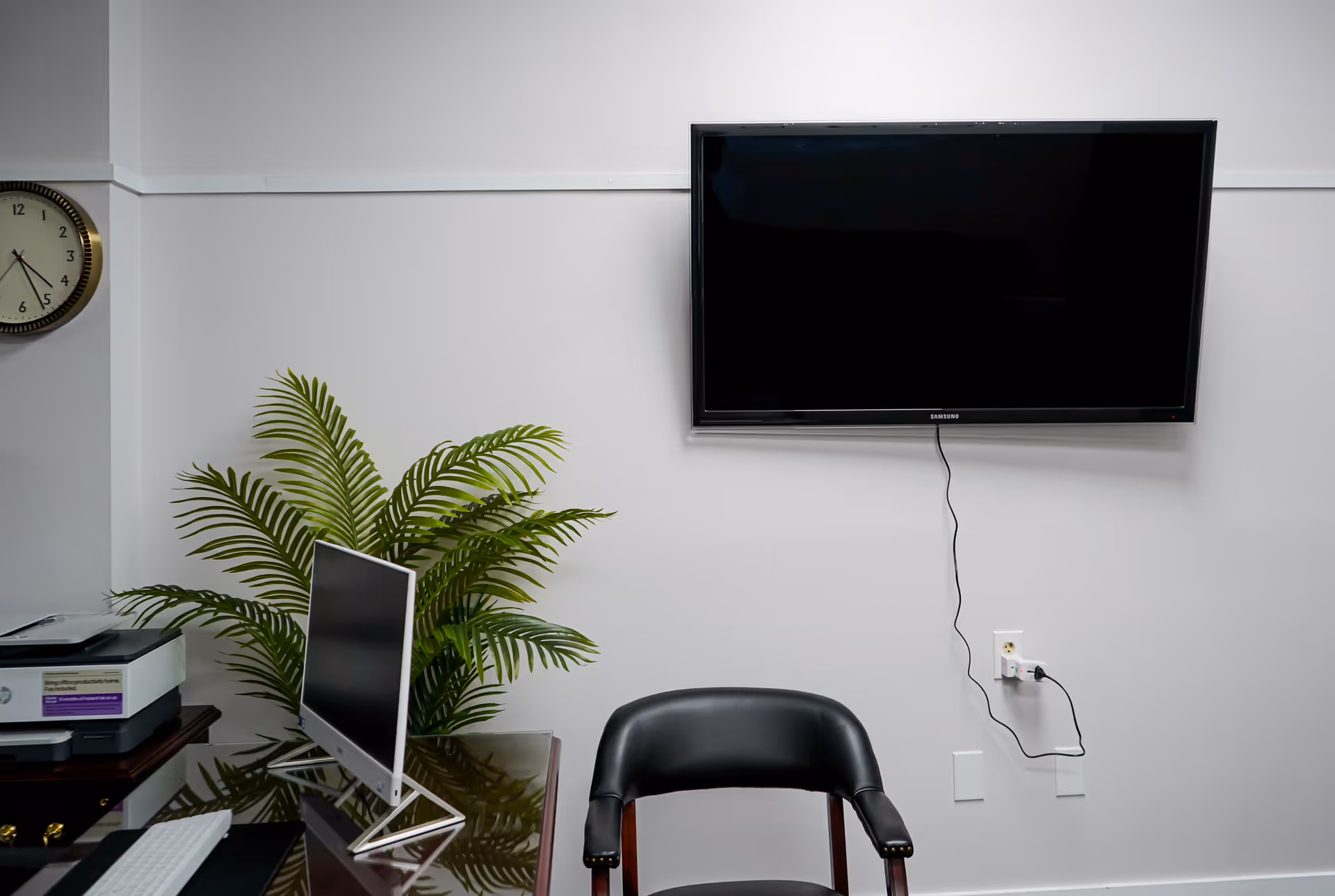 Office area with a desk, monitor, printer, plant, clock, and wall-mounted TV.