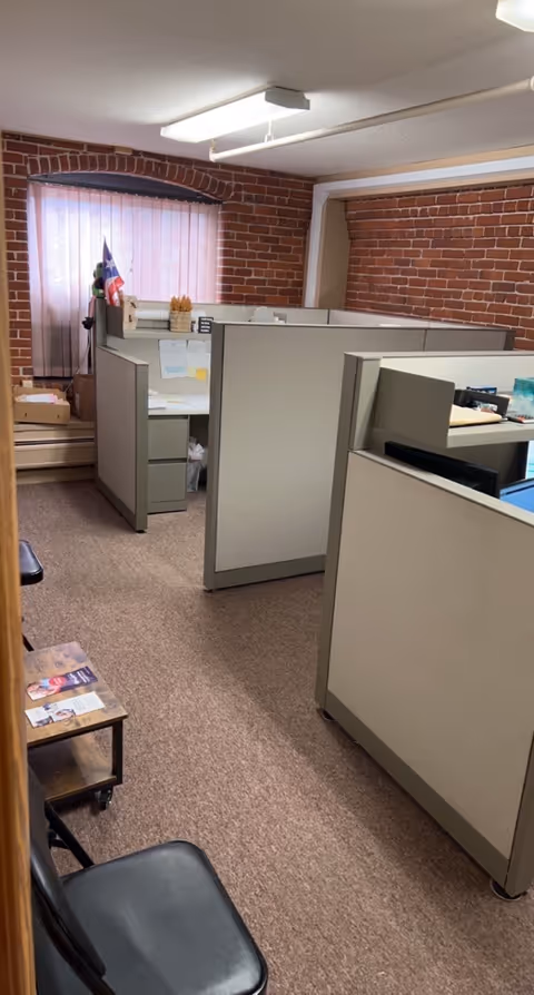 Office cubicles inside a brick-walled workspace with papers and a small flag.