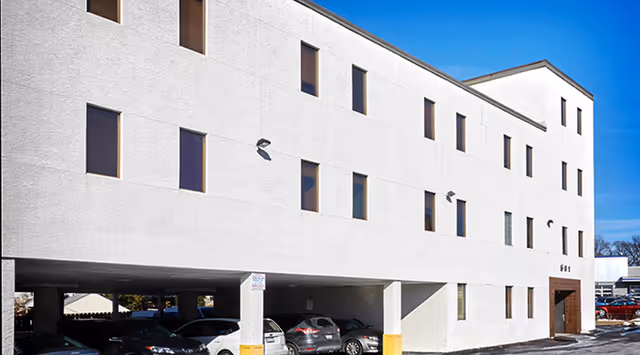 White multi-story senior living building with parked cars beneath and a blue sky.