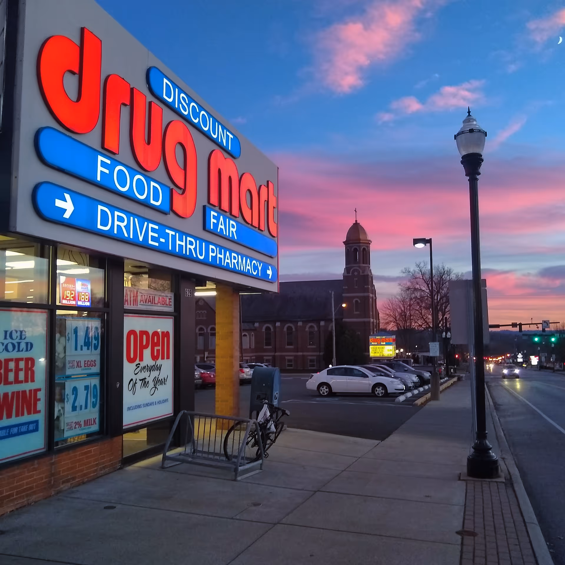 Storefront of Discount Drug Mart with large neon sign and drive-thru pharmacy at dusk.