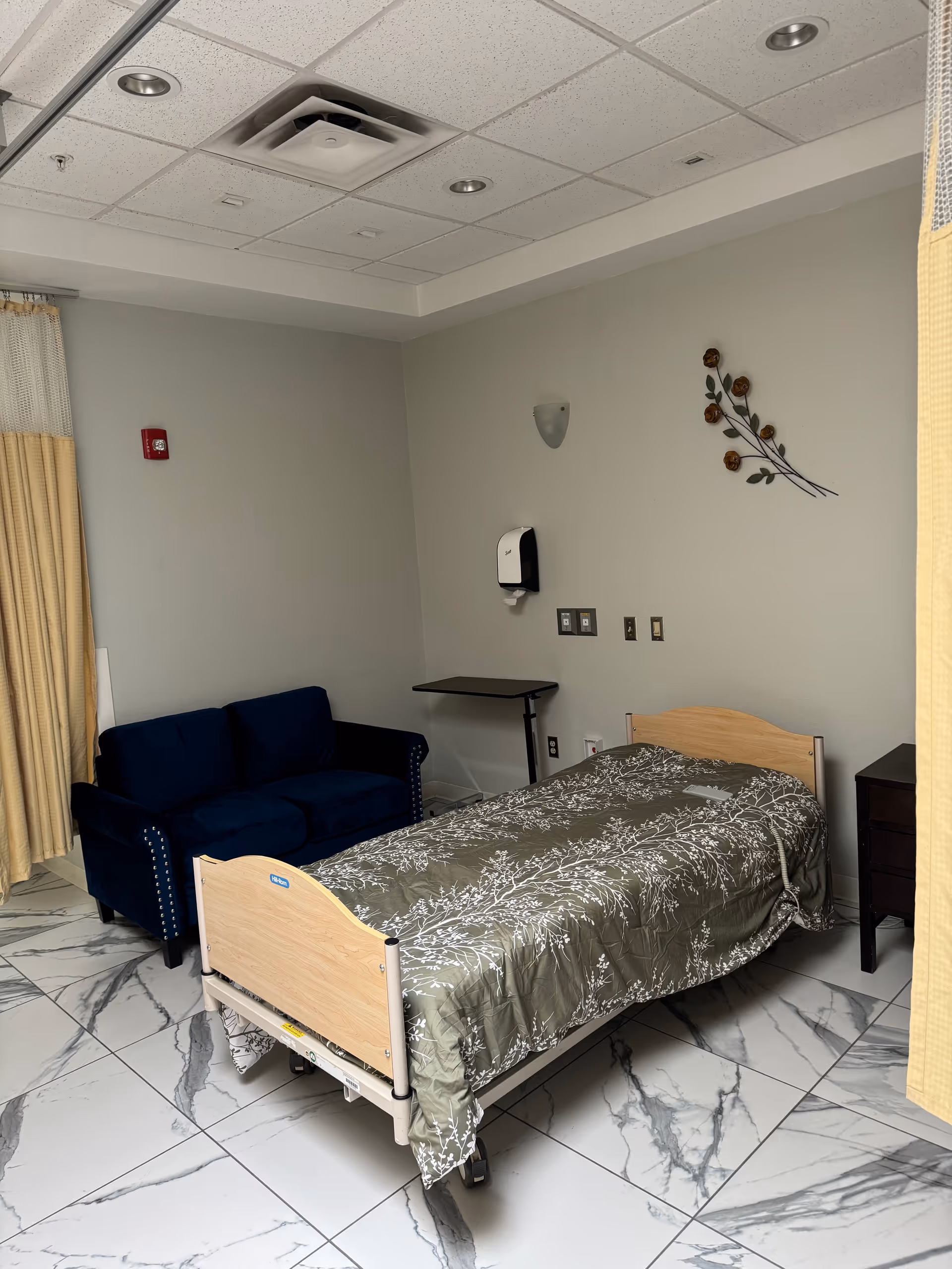 A patient bedroom in a senior facility with a bed, blue couch, and tiled floor.