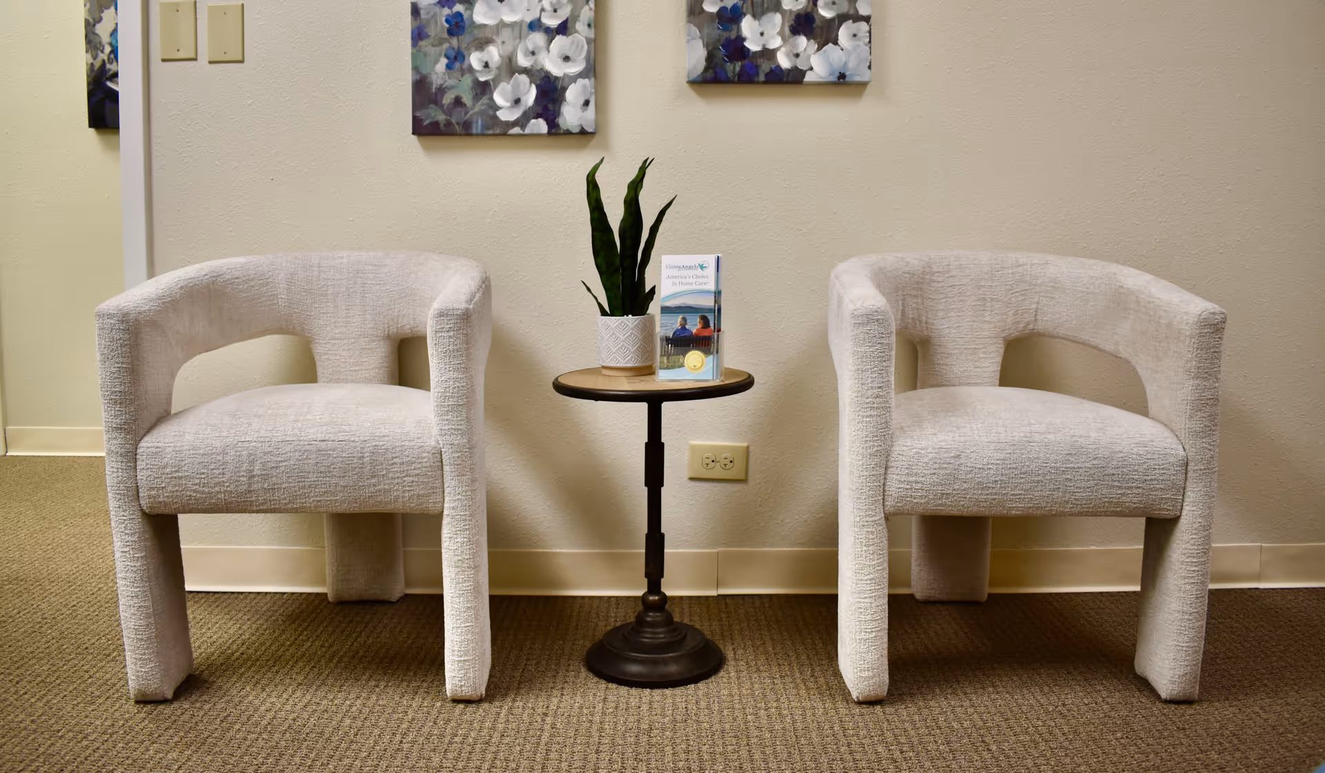 Two light-grey chairs flank a small round table with a plant and brochures.