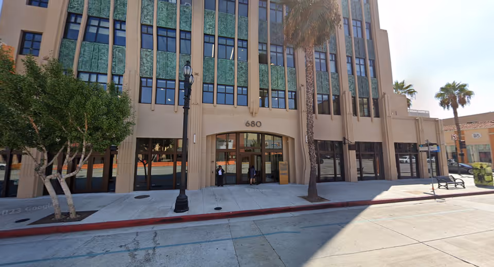 Beige, multi-story building facade with glass doors, street sidewalk, and palm trees.