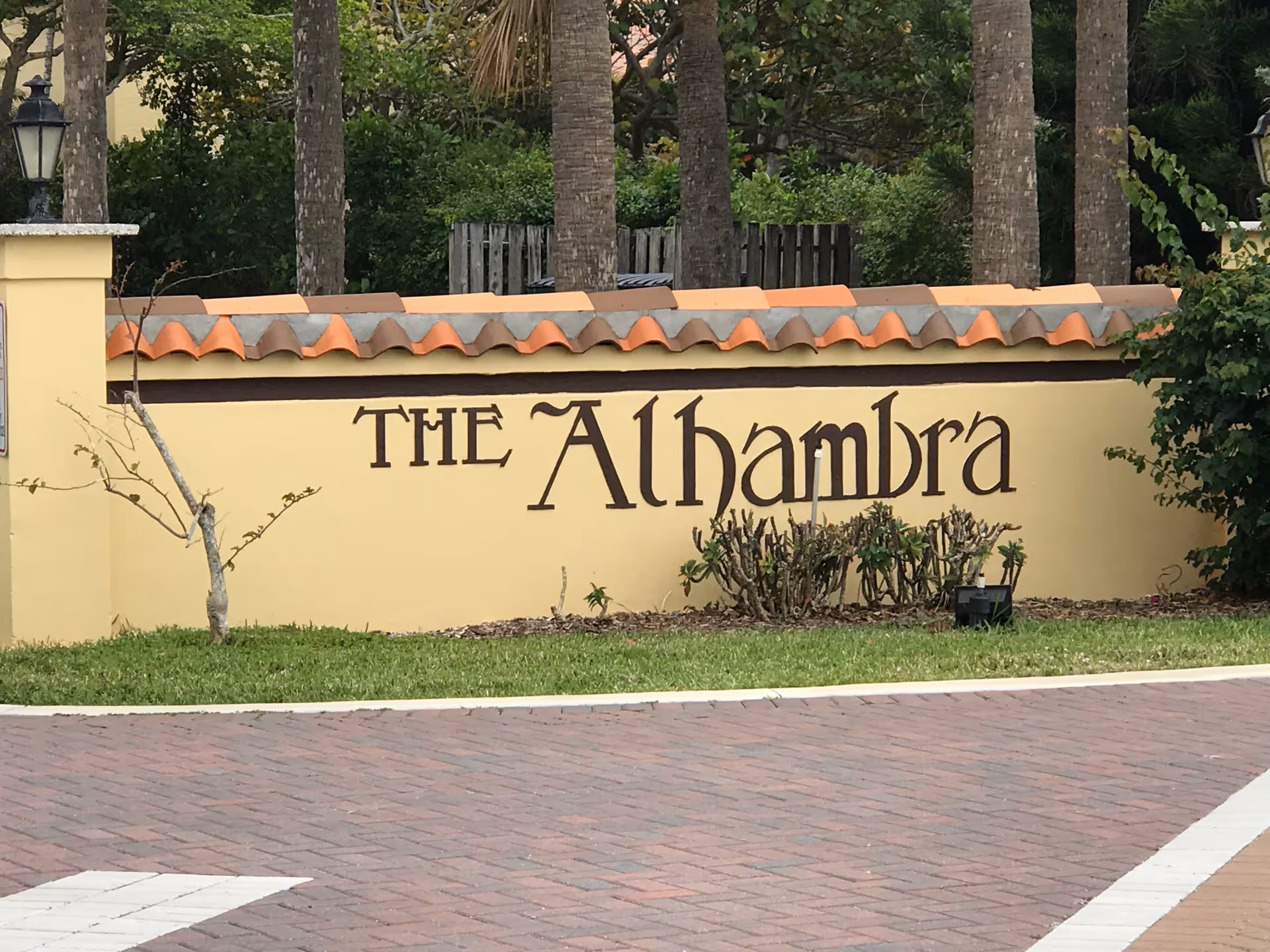 Sunlit entrance wall with 'The Alhambra' sign, palm trees, and landscaped shrubbery.