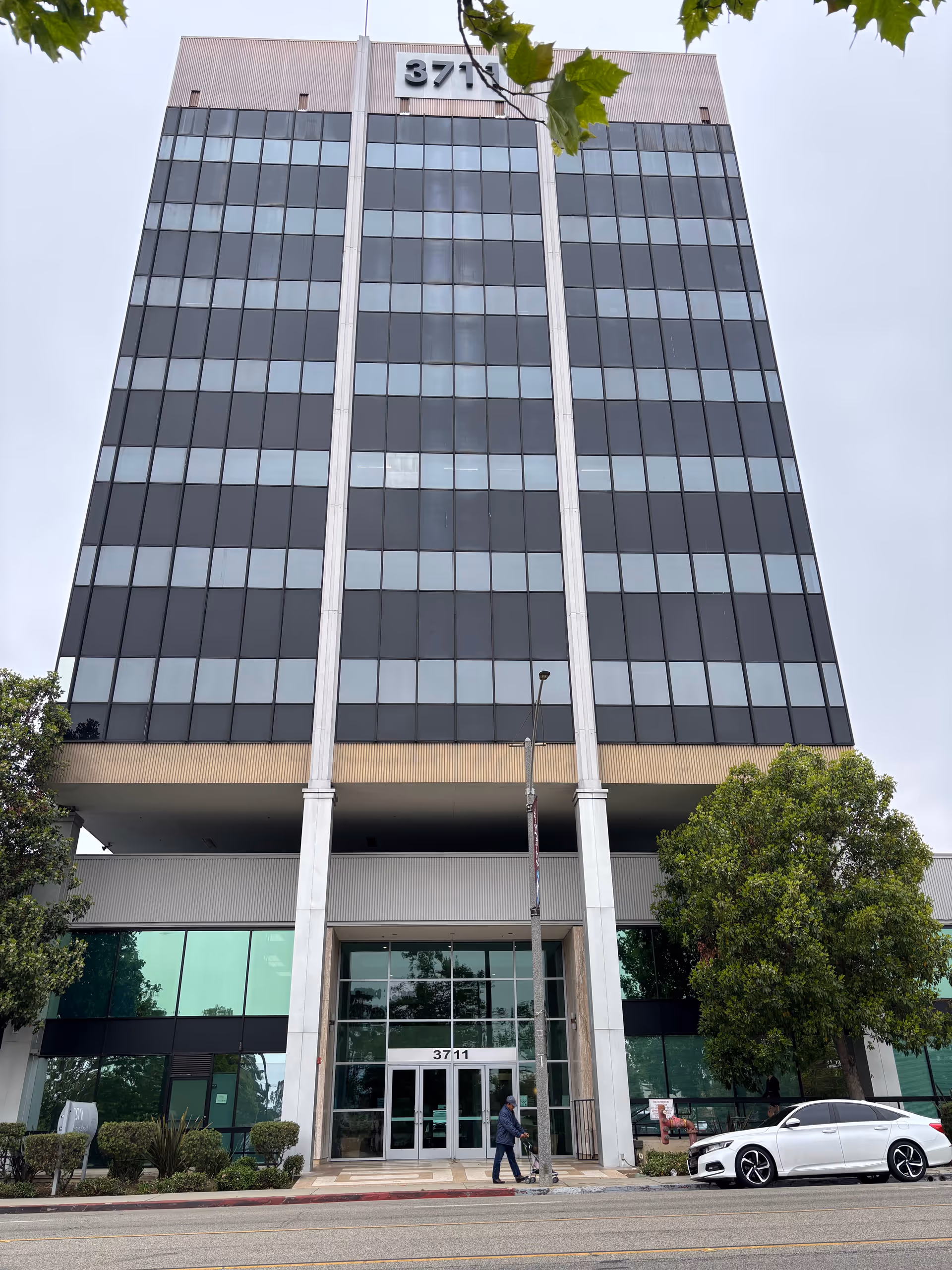 Tall glass-front office building with main entrance, street, trees, and a person walking.