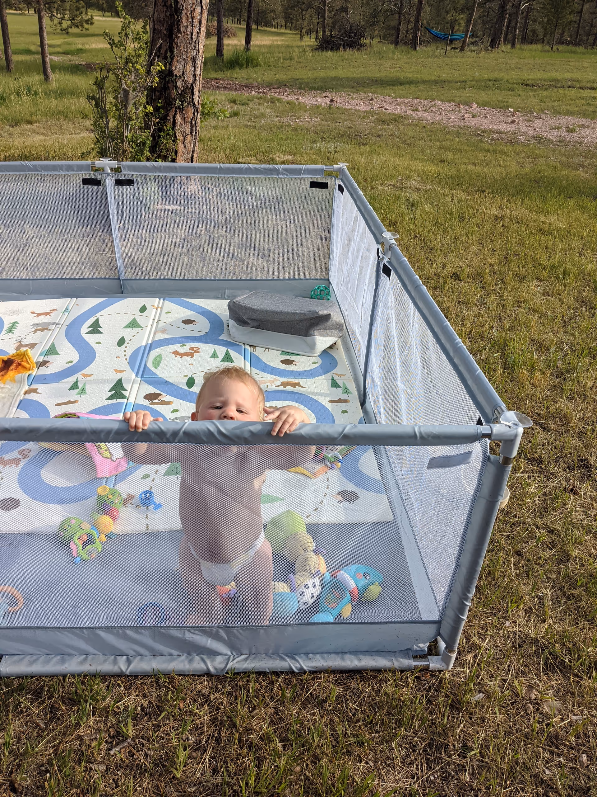 Baby standing inside a mesh outdoor playpen with colorful toys on a grassy yard.