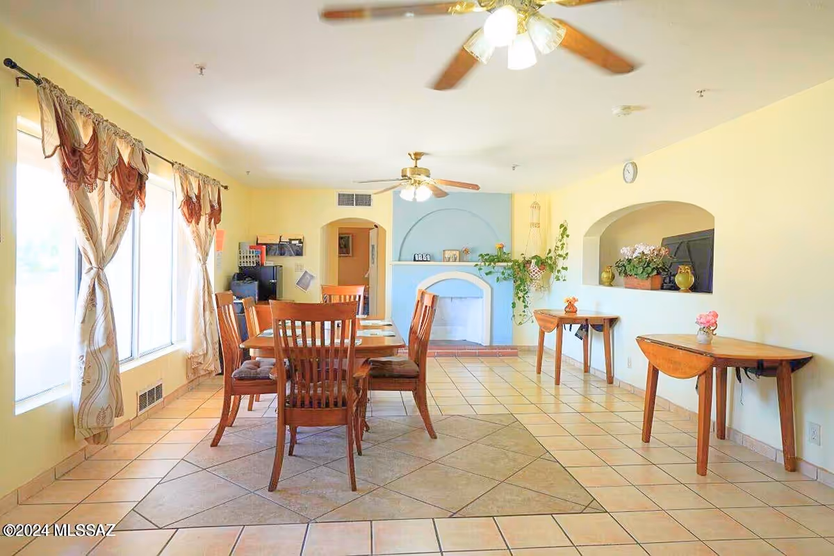 Sunlit dining room with wooden table and chairs, windows, pastel yellow walls, fans, and plants.