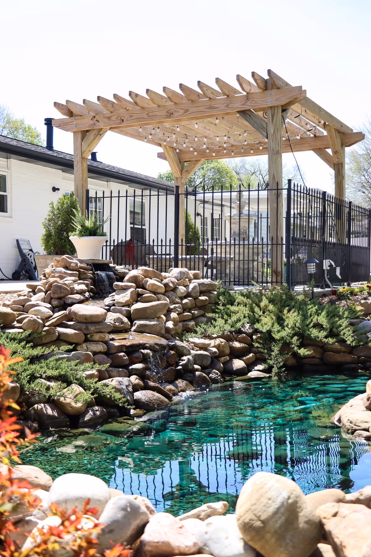 Outdoor communal space with a wooden pergola, string lights, rock waterfall, and clear pool.