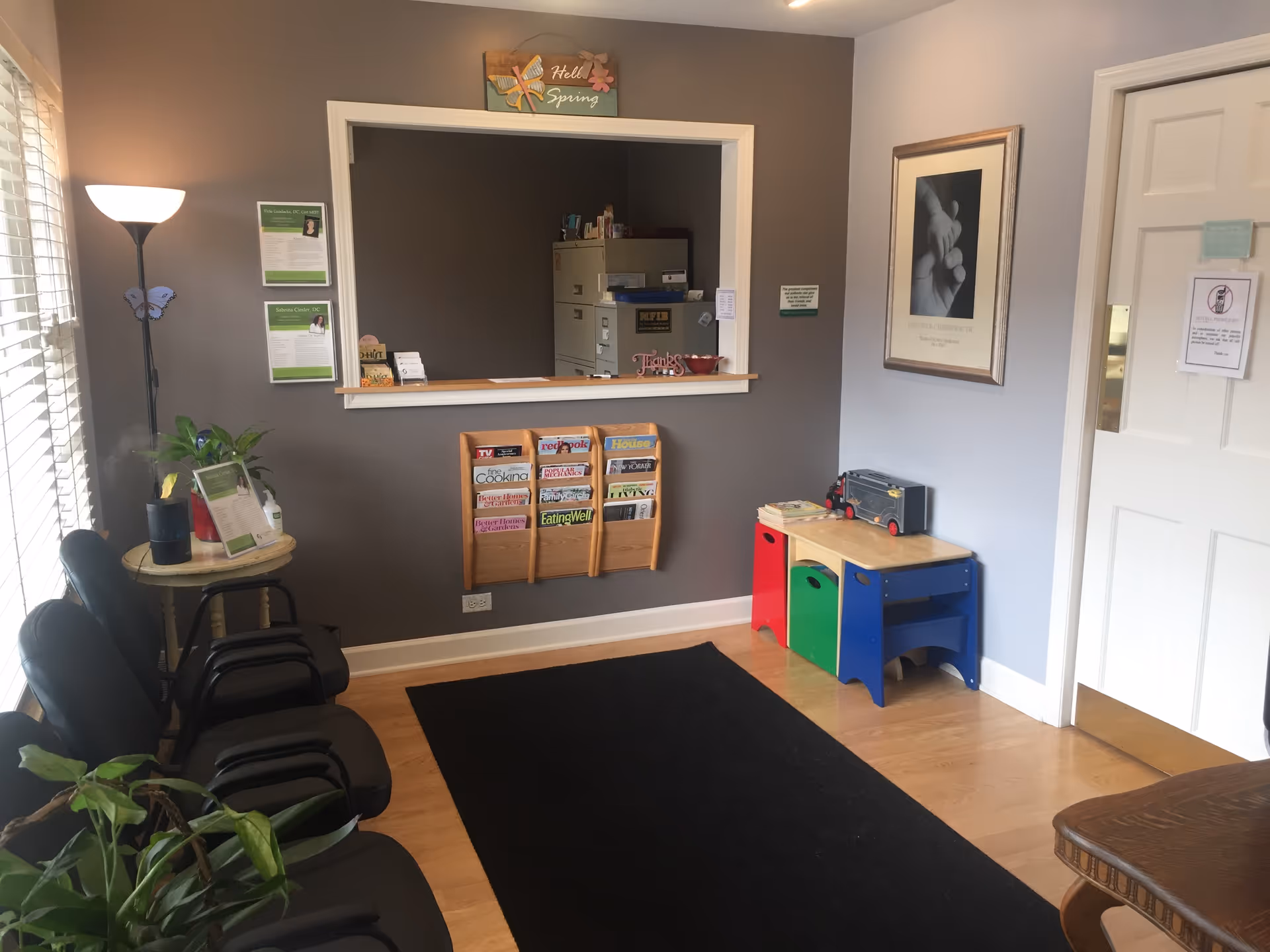 Waiting area with chairs, a magazine rack, plants, and a windowed reception pass-through.