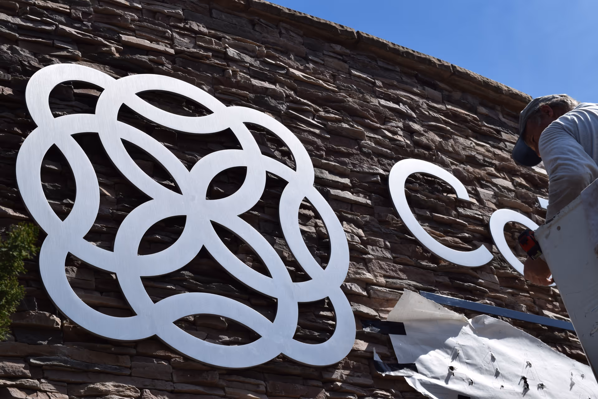 A worker installs a large white abstract sculpture on a stone building exterior.
