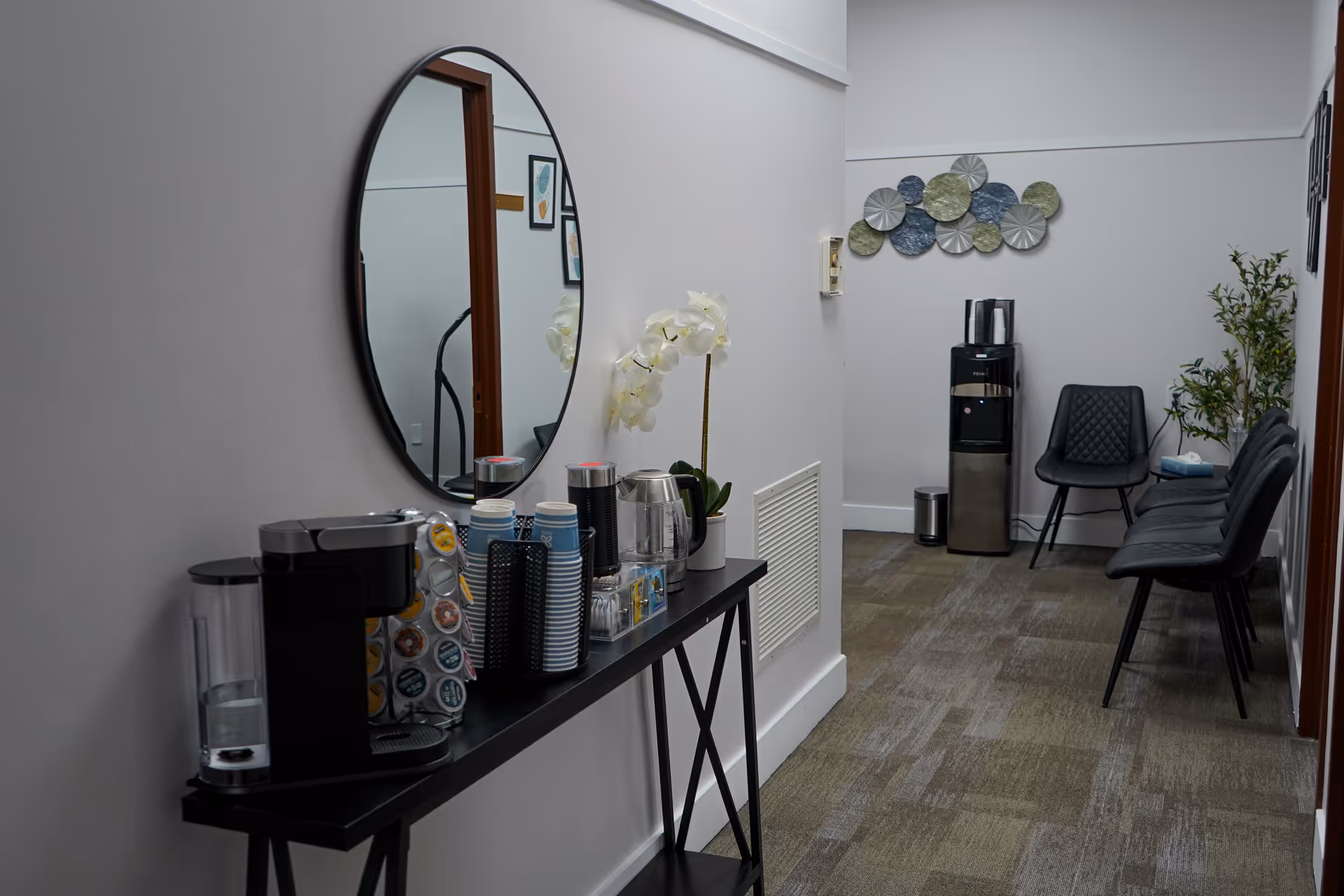 Coffee station with cups and a round mirror along a hallway in a senior facility lounge.