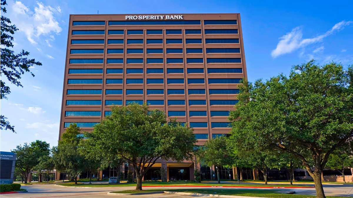 Tall brick bank building labeled Prosperity Bank with green trees and a clear blue sky.