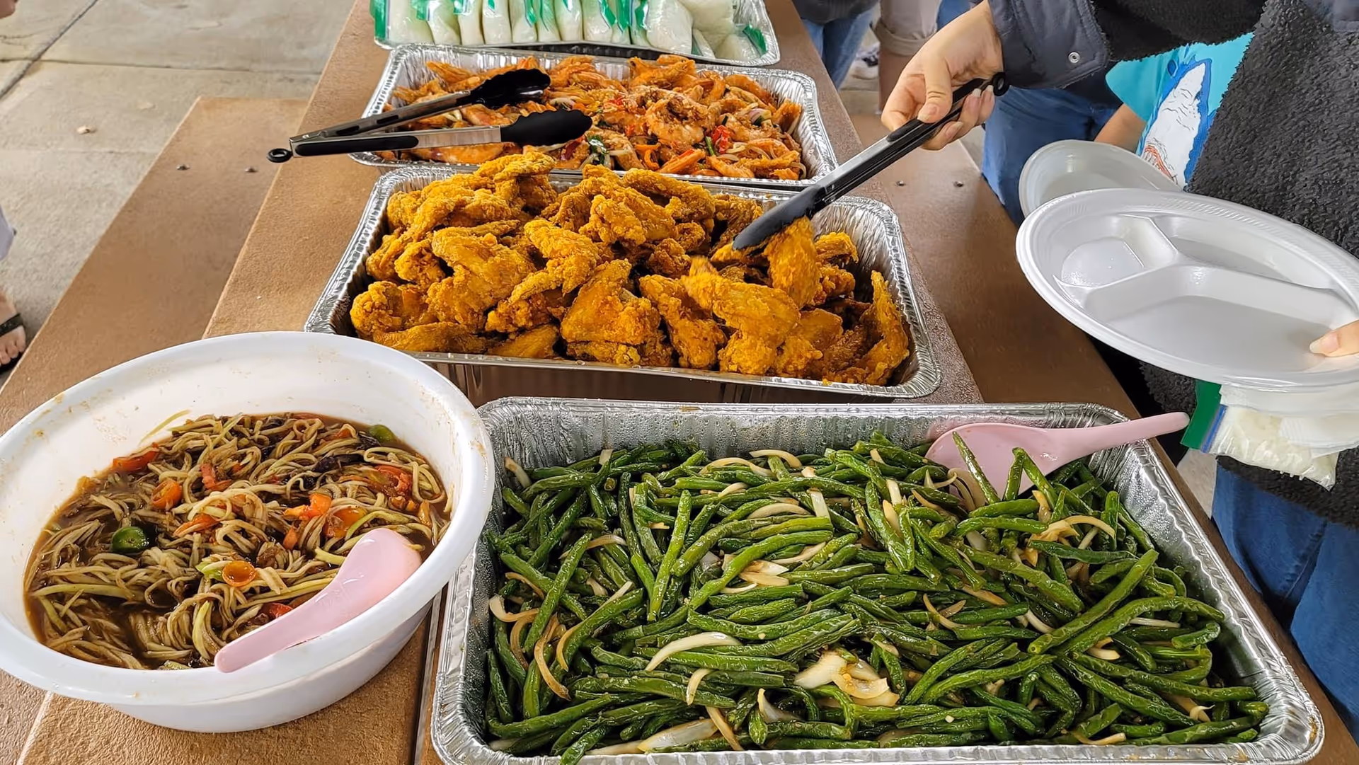 Buffet table displaying fried chicken, green beans, and noodles with serving tongs and plastic plates.