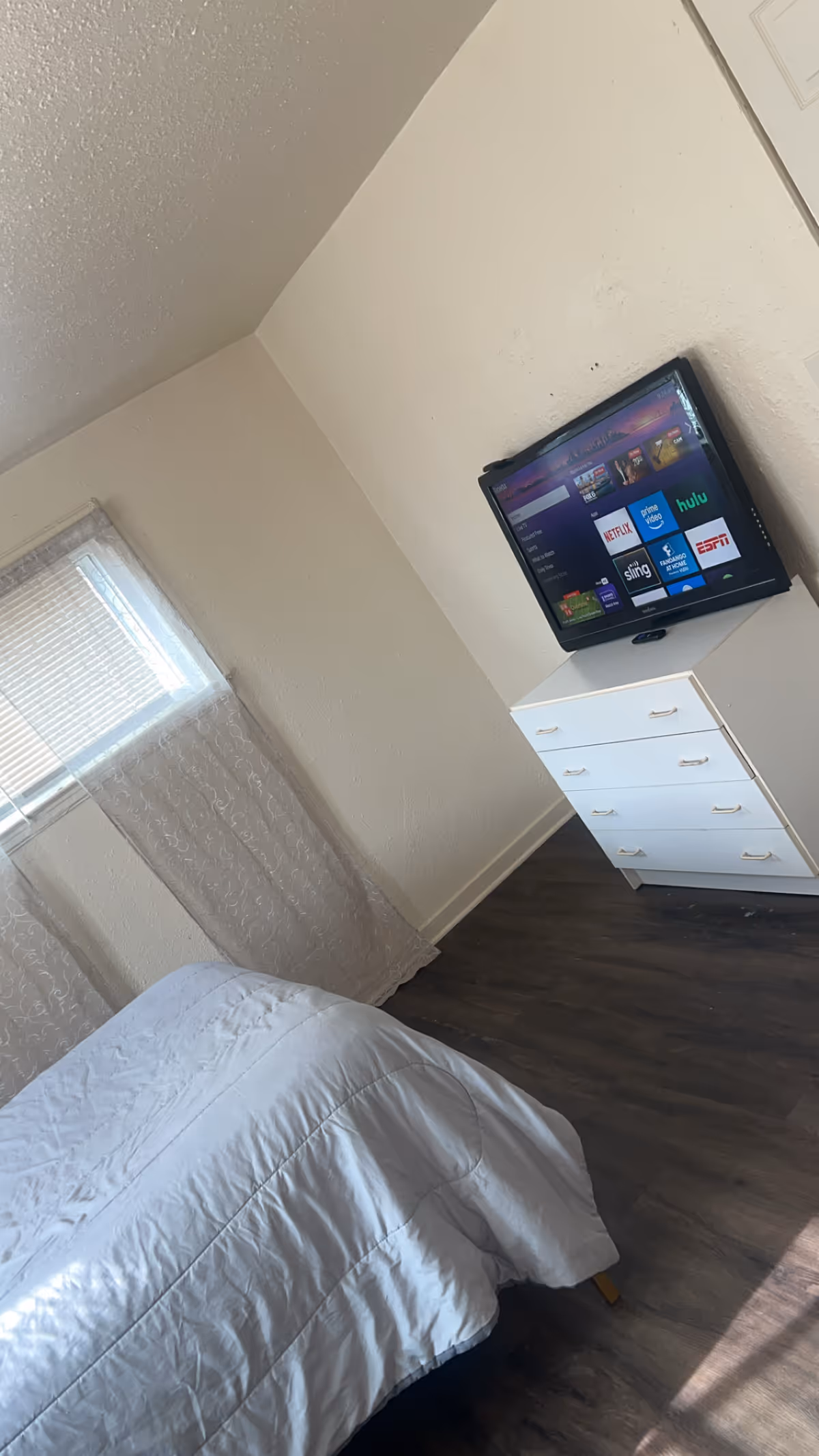 A bedroom with a bed, window curtains, and a wall-mounted TV above a white dresser.