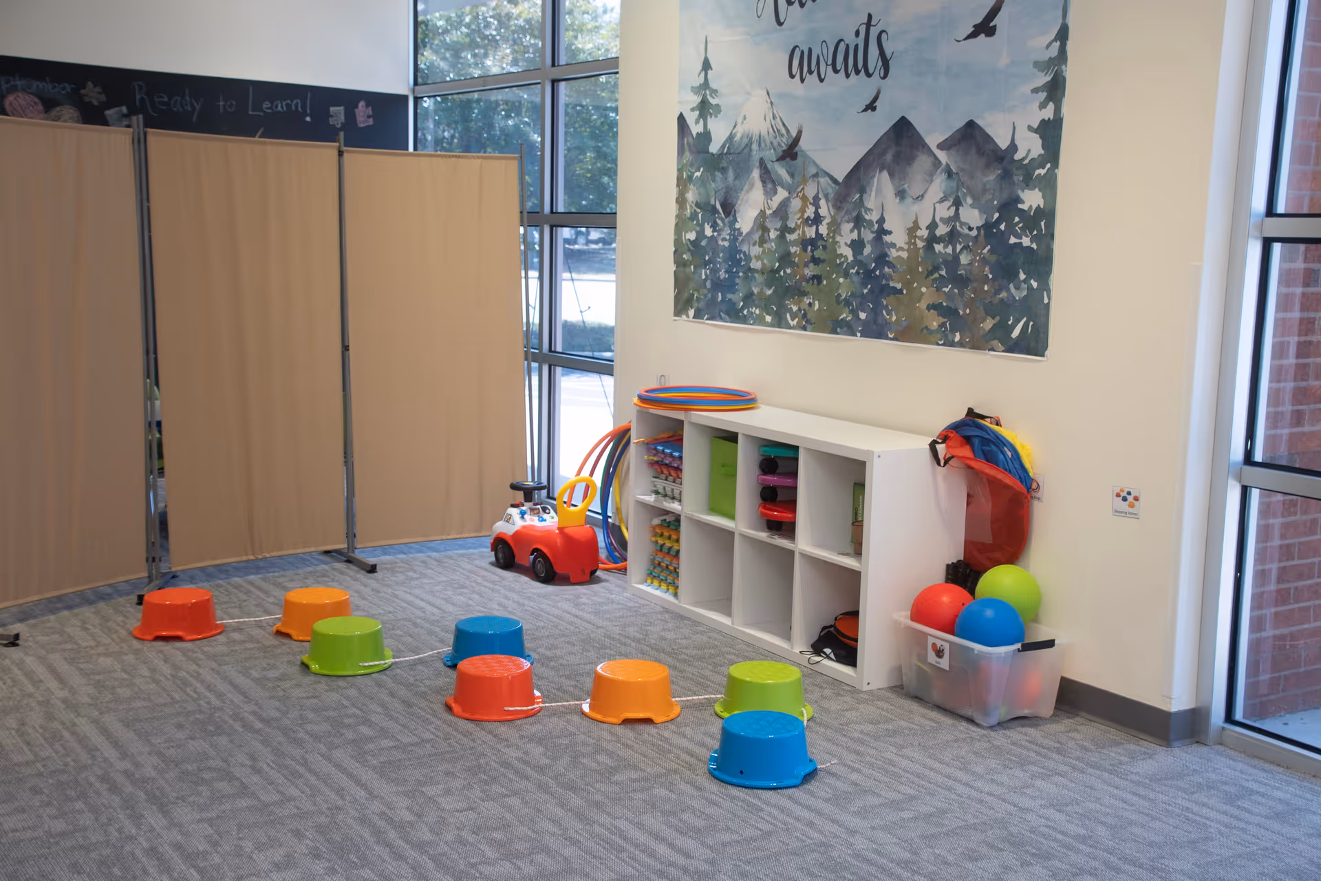 Bright indoor playroom with colorful stools, toys, cubby shelves, and partition screens.