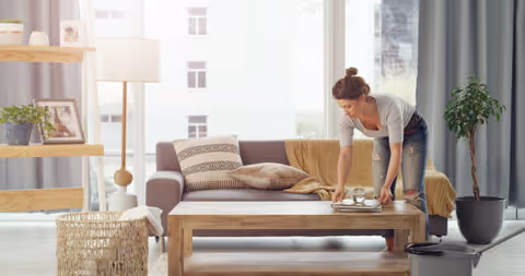 A caregiver tidies a cozy living room with sofa, coffee table, and plants.