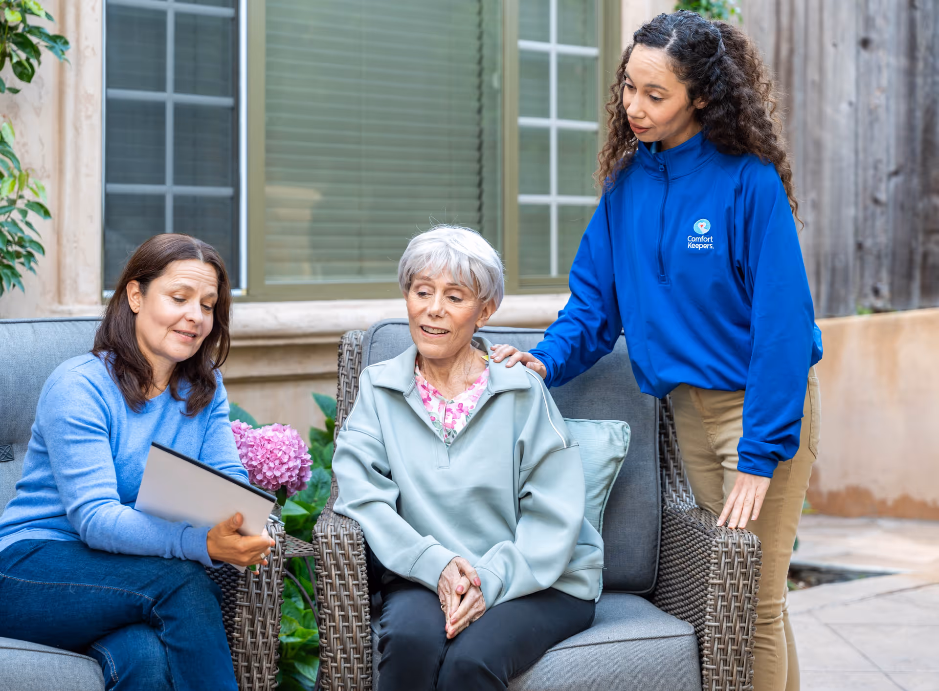 Outdoor patio with a caregiver and two residents seated on wicker furniture.