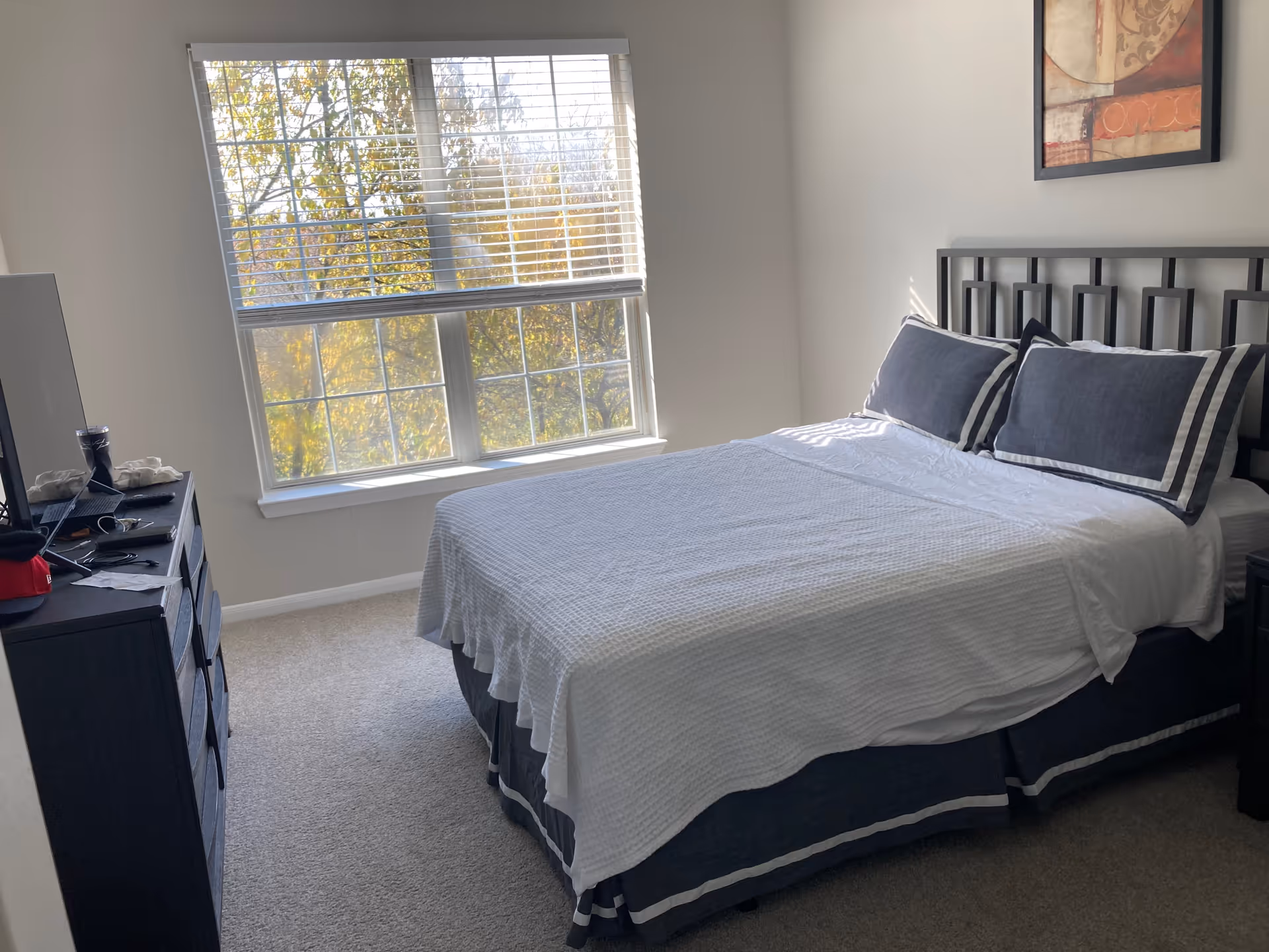 Neatly made bedroom with a queen bed, dark dresser, and large window overlooking trees.