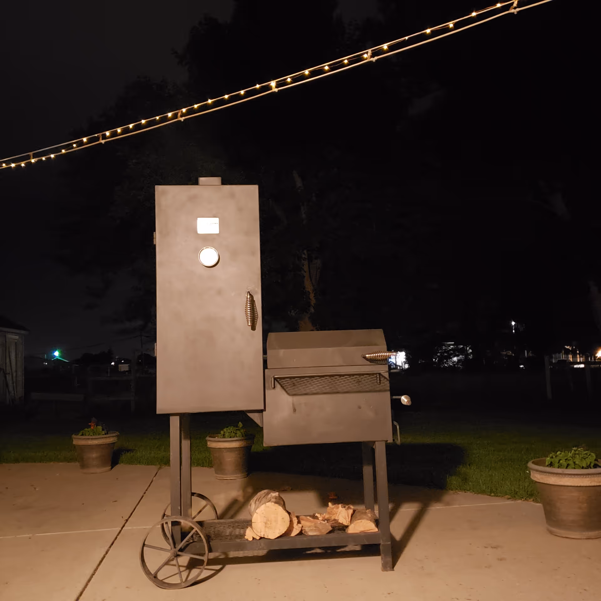 Nighttime outdoor scene with a metal barbecue grill on wheels, string lights above, and potted plants.