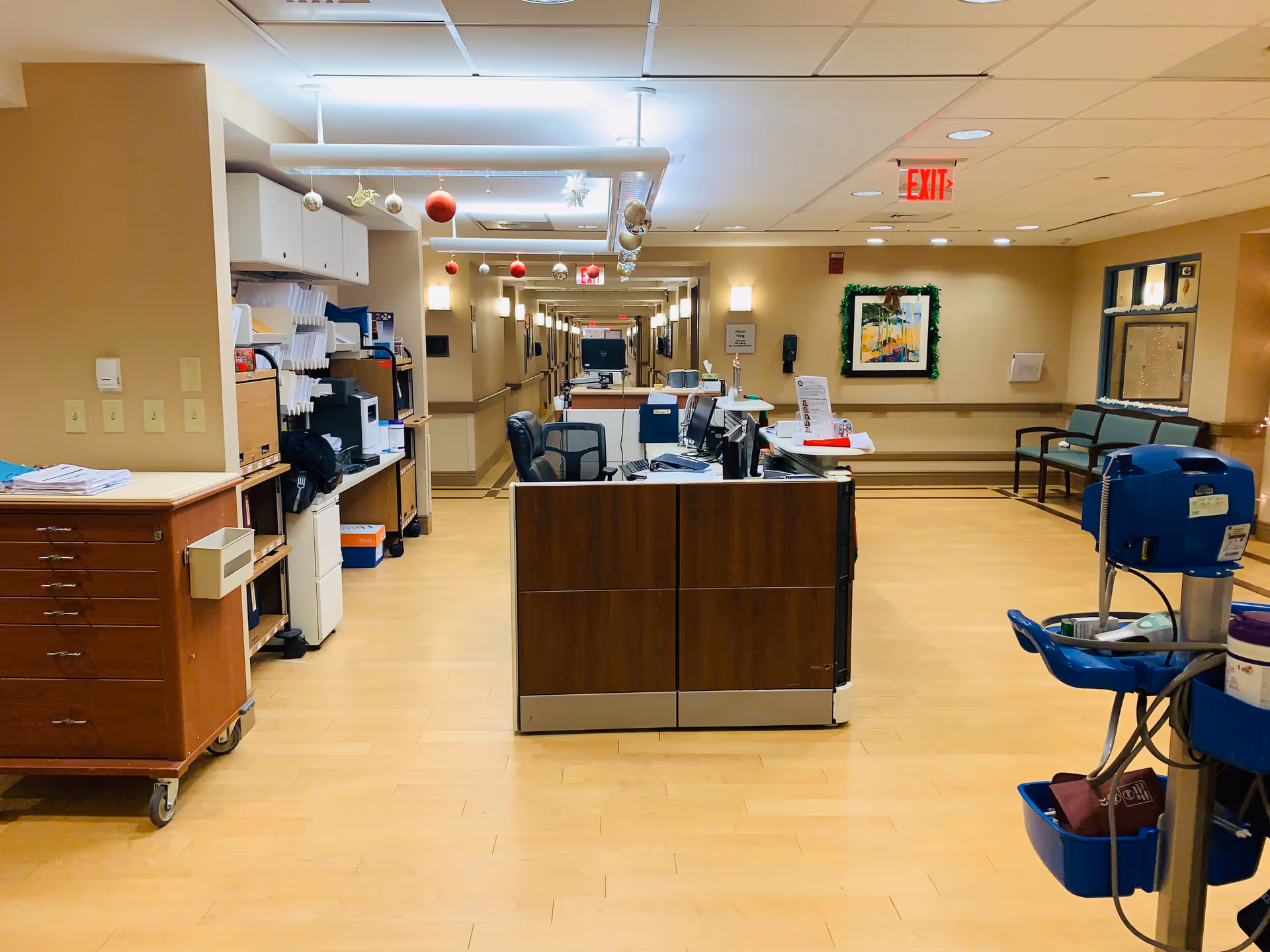 Nursing station with desks, medical equipment, and a well-lit hallway in a senior living facility.