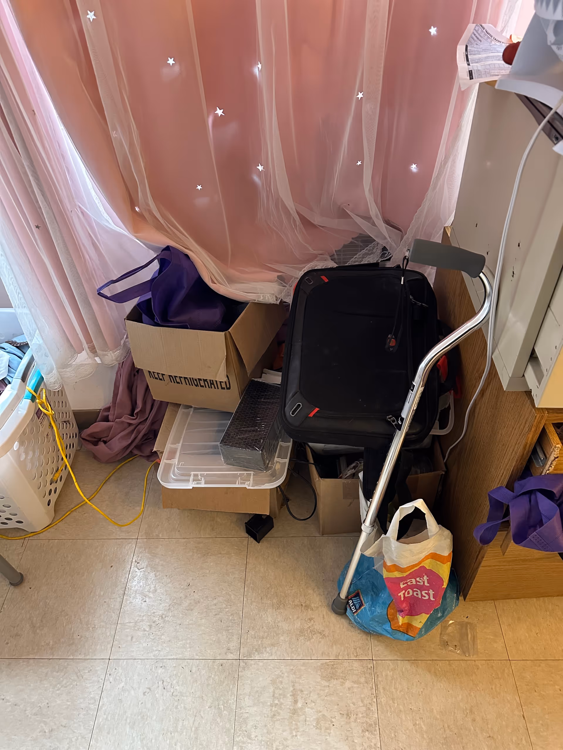 Cluttered interior corner with pink sheer curtain, walker, boxes, and bags.