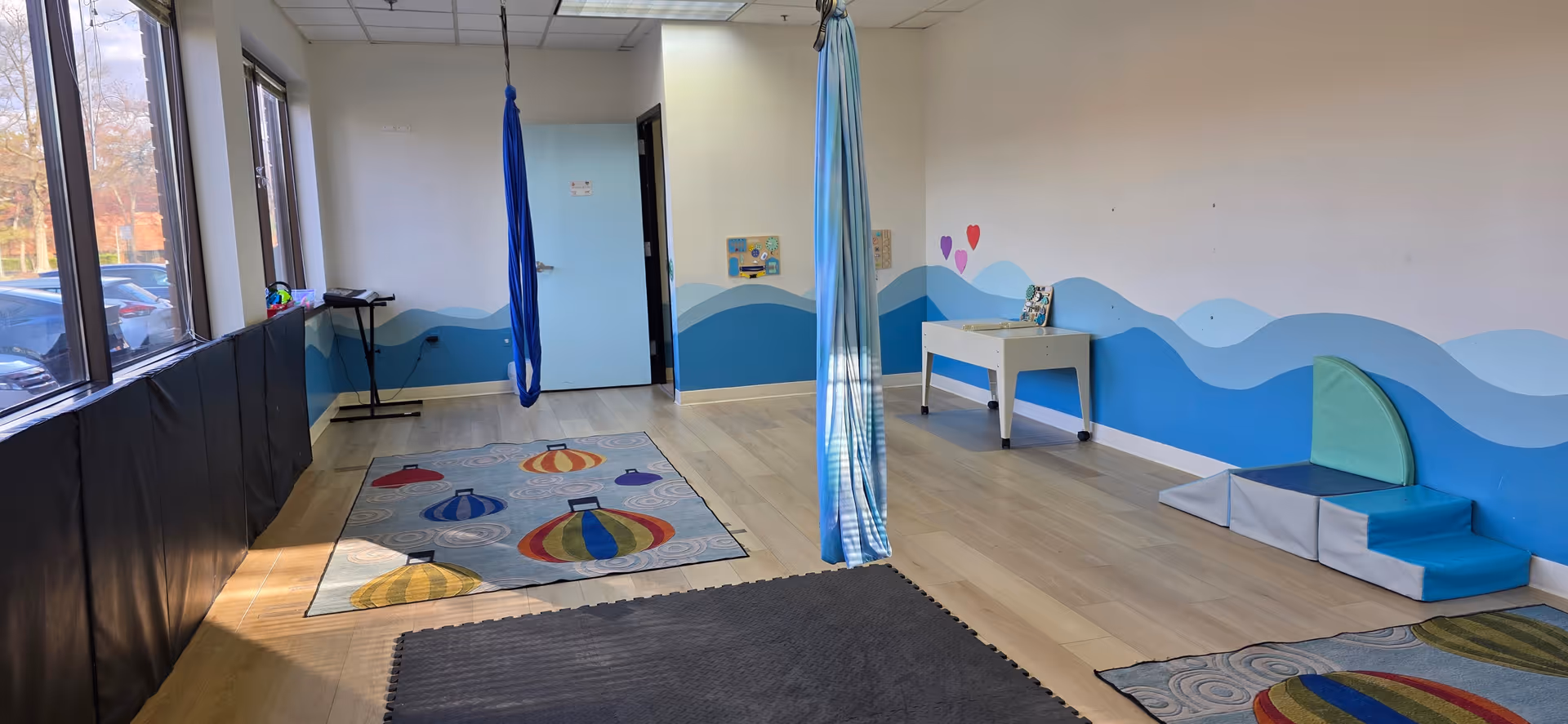 Bright therapy room with blue wavy mural, mats, play equipment, and aerial silks.