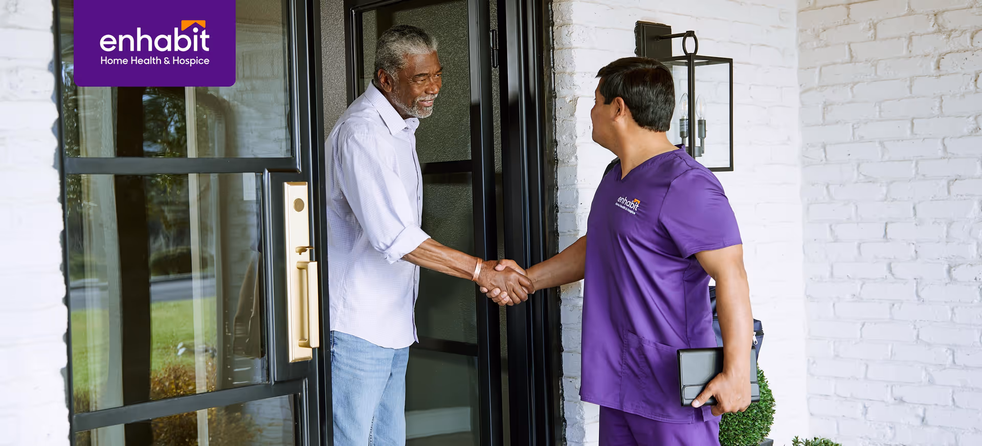 Two people shake hands at a building entrance; one wears purple scrubs with Enhabit logo.