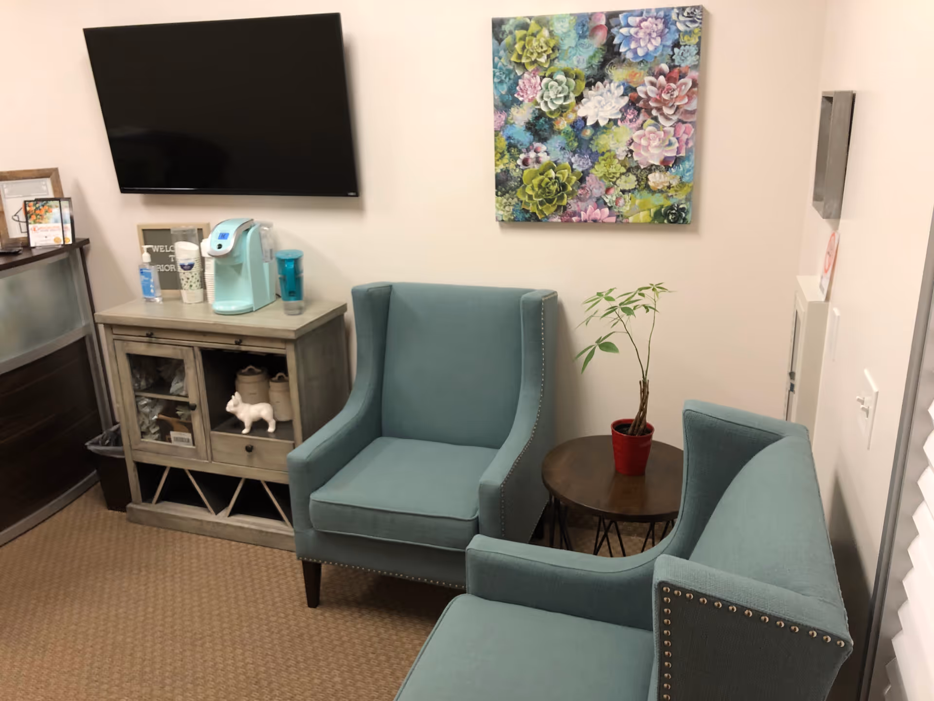 Small teal living room lounge with two chairs, a coffee table, plant, wall art, and TV.