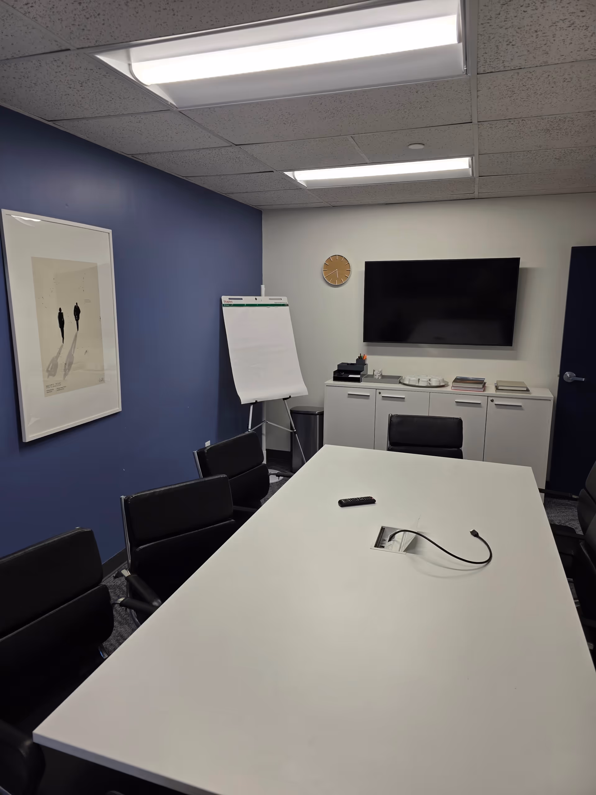 Modern conference room with a long table, black chairs, cabinets, and a wall-mounted TV.