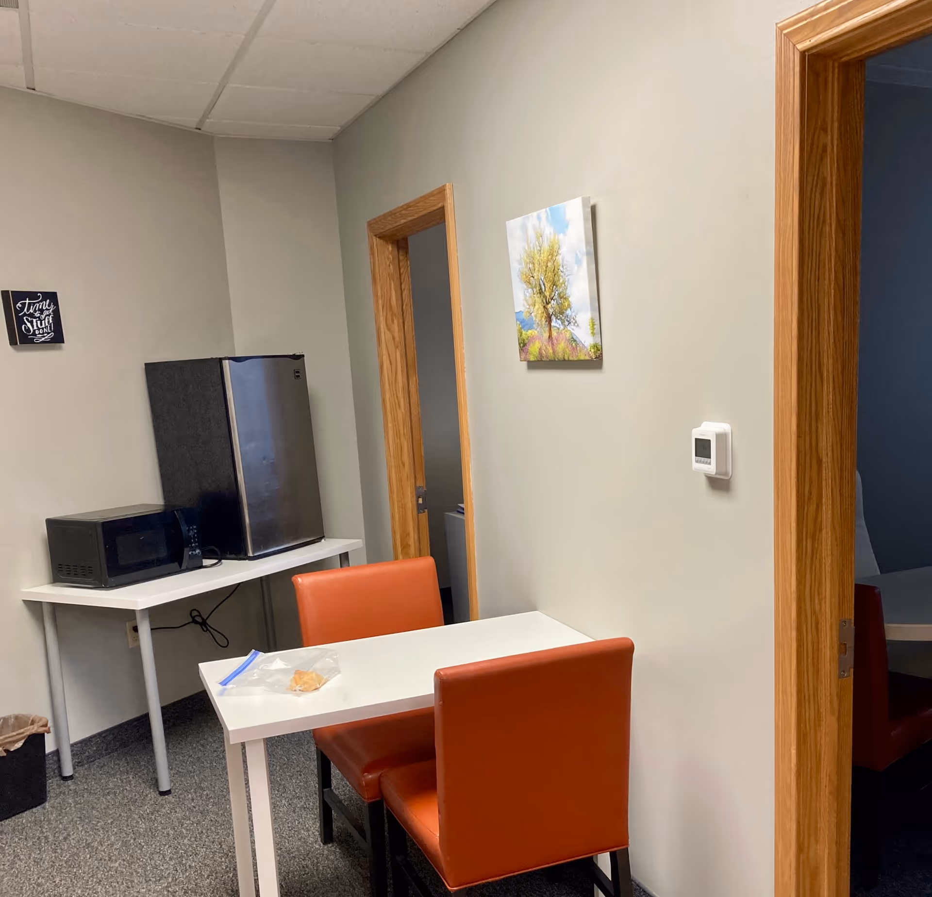 Small staff break room with microwave, fridge, table, orange chairs, and wall art.