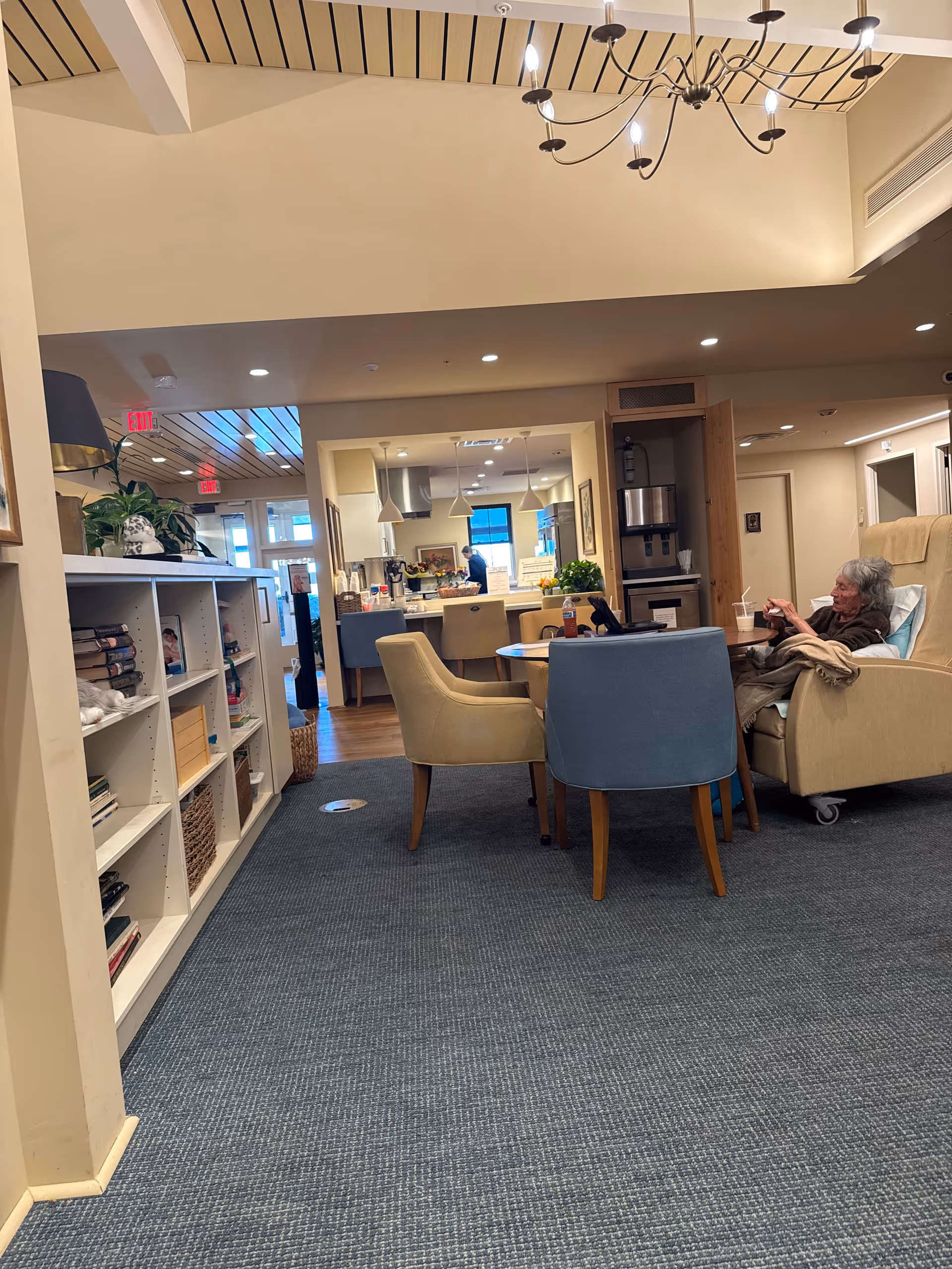 Lounge area with blue and beige chairs, a bookshelf, and an elderly resident resting.