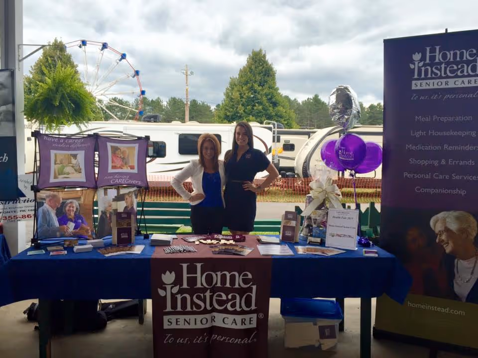Two women stand behind a Home Instead promotional booth outdoors with banners and brochures.