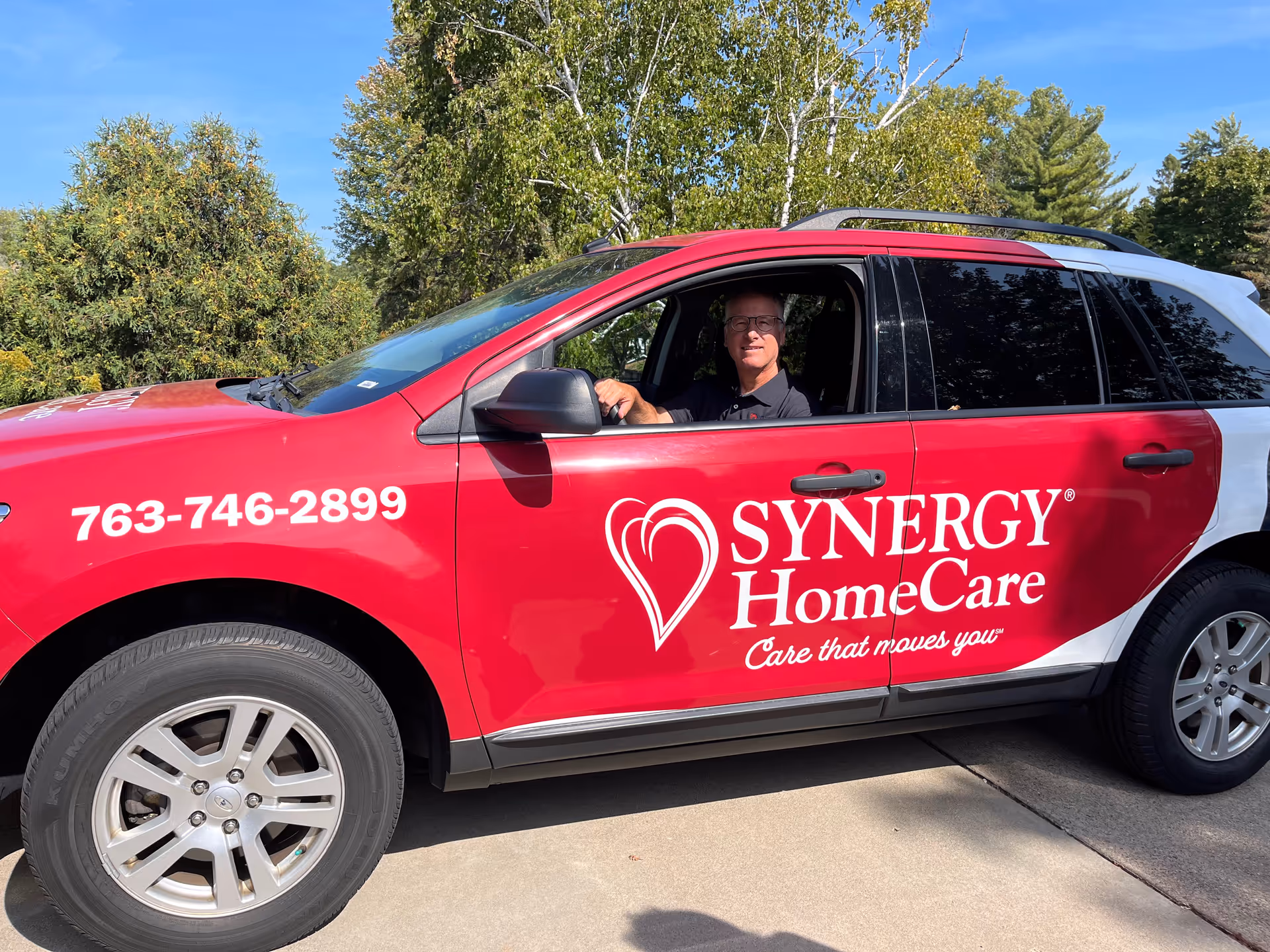 Red SYNERGY HomeCare van parked outdoors with driver inside, logo and heart emblem visible.