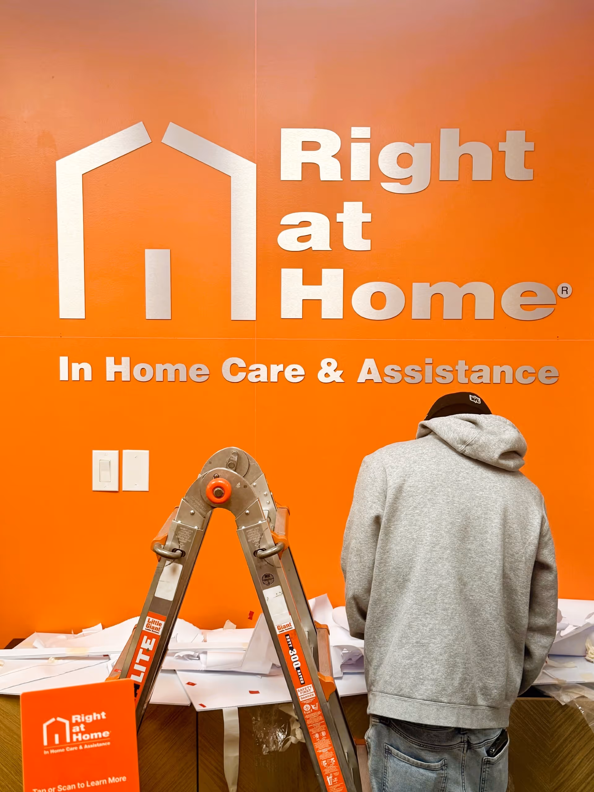 Bright orange wall displays Right at Home branding, with a person in gray hoodie near ladder.