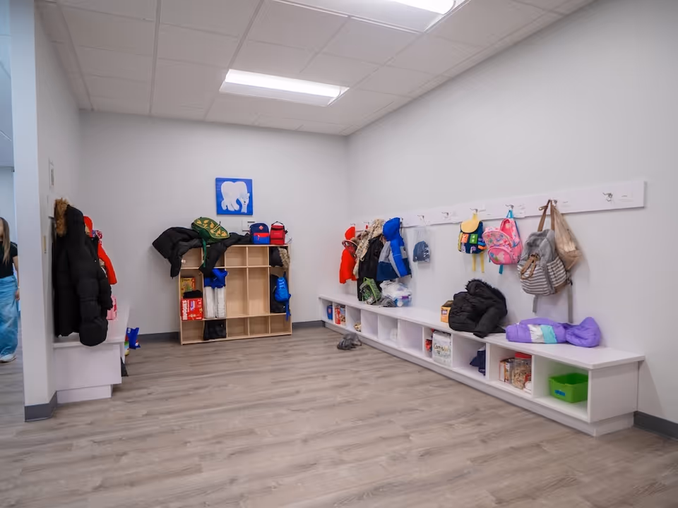 Bright interior coatroom with cubbies, hooks, coats, and backpacks along white walls.