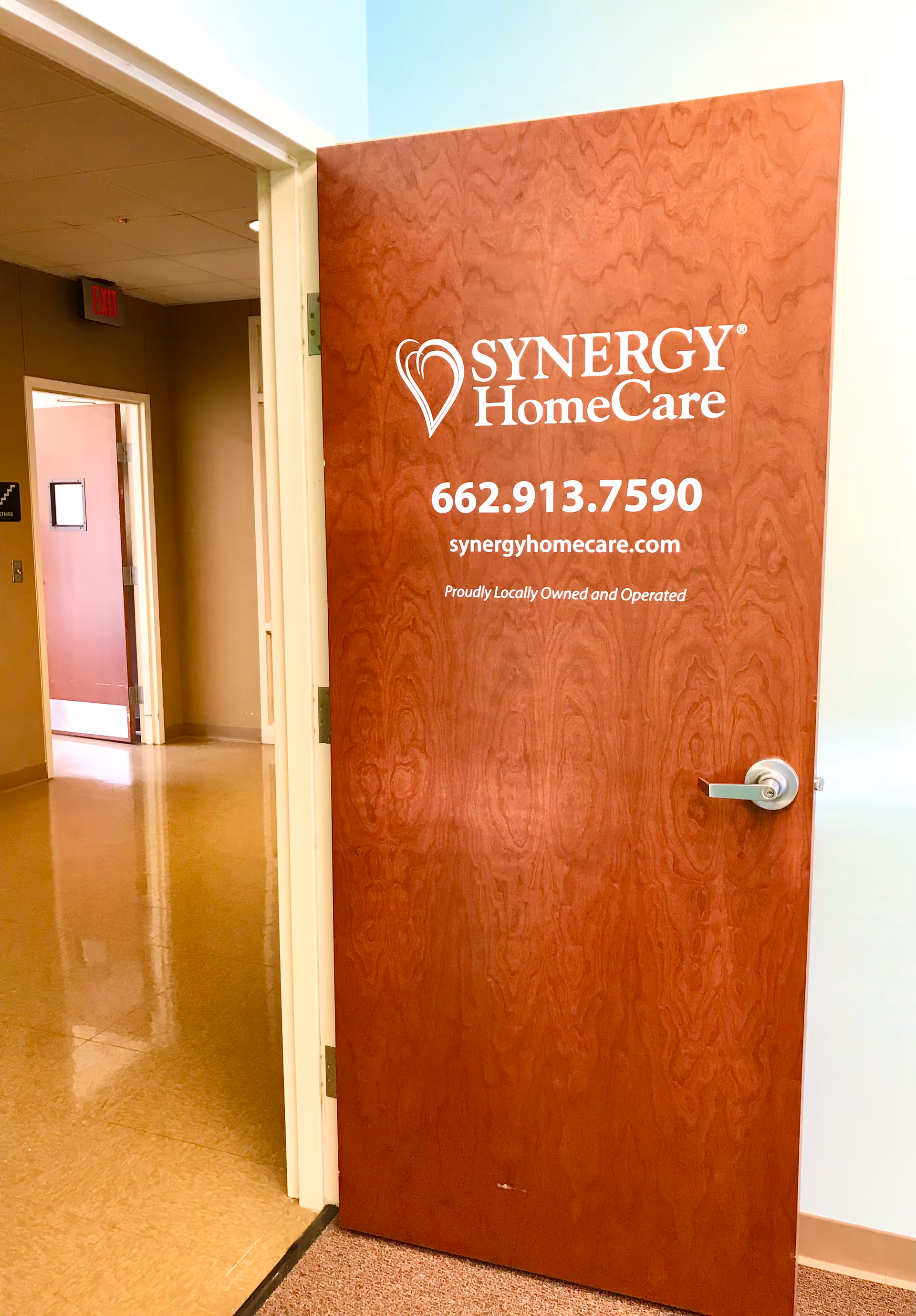Open doorway with Synergy HomeCare sign and contact info on wooden door in hallway.