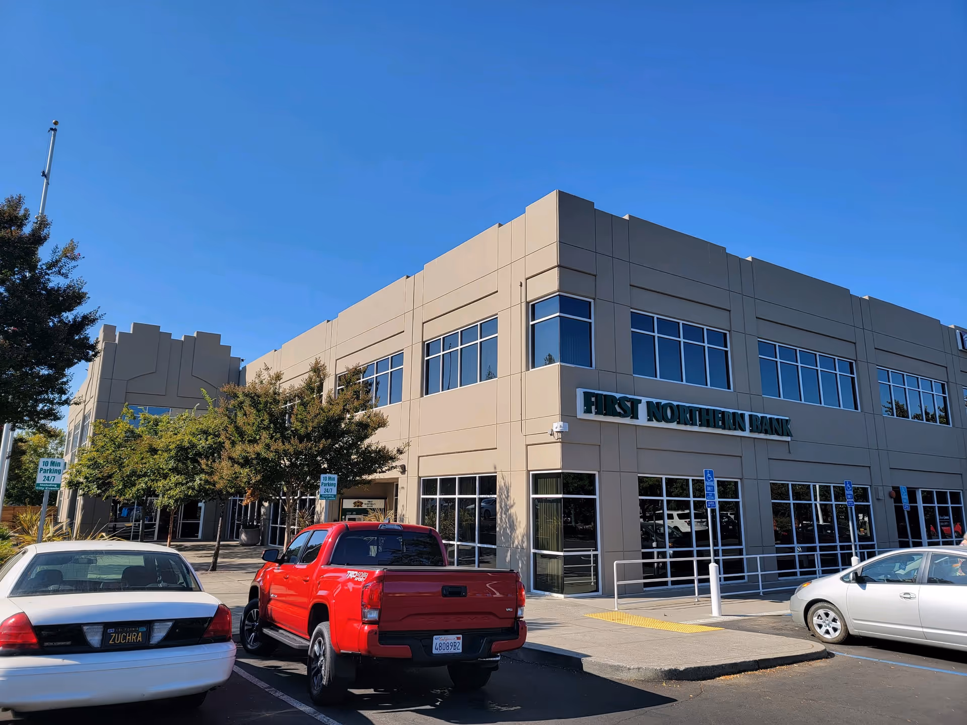 Two-story beige bank building with large windows and cars parked outside.
