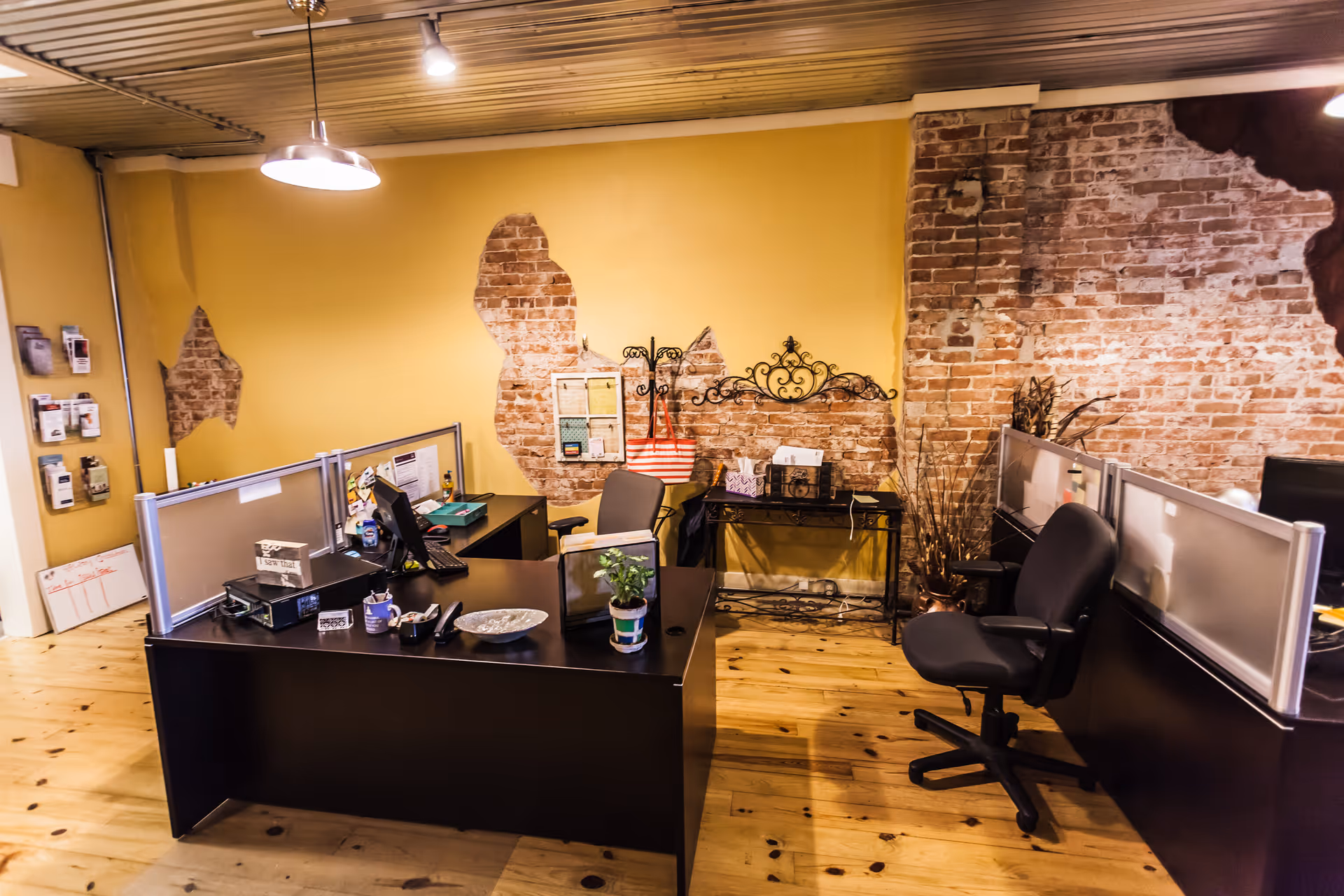 Office workspace with desks, cubicles, chairs, and exposed brick wall in warm lighting.