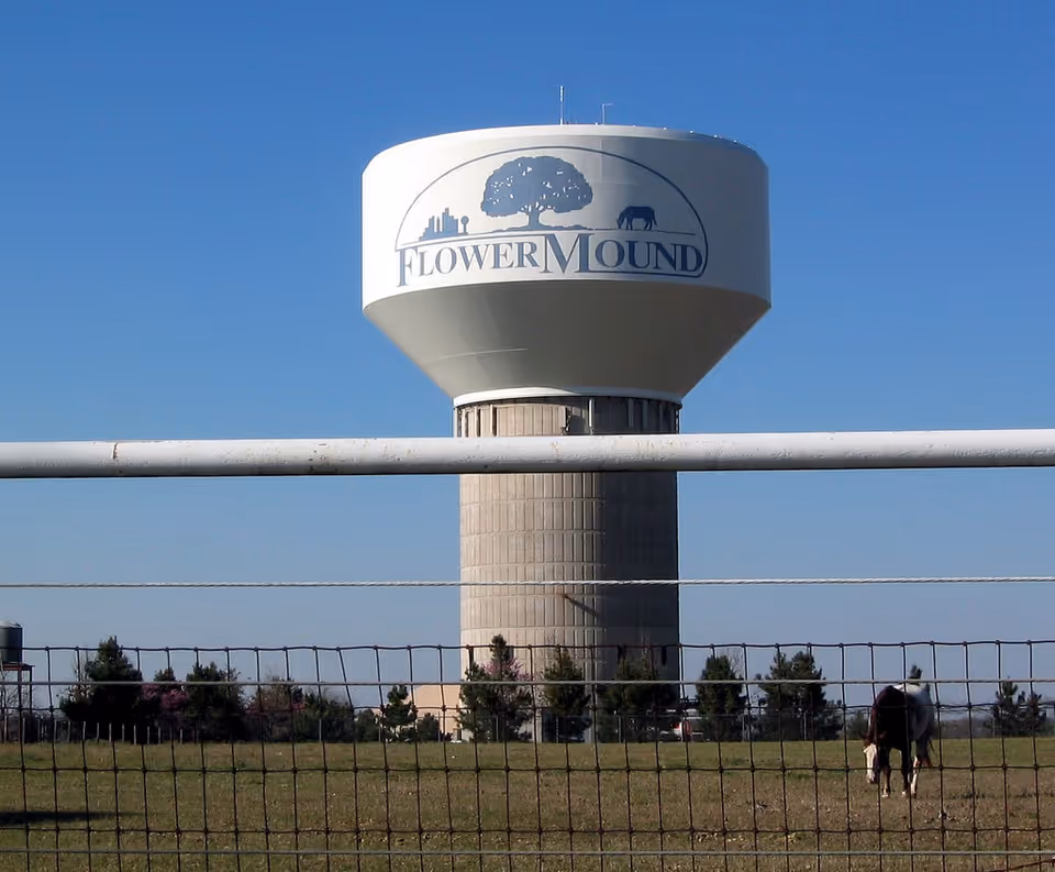 Water tower with Flower Mound logo rises above a fenced grassy field.