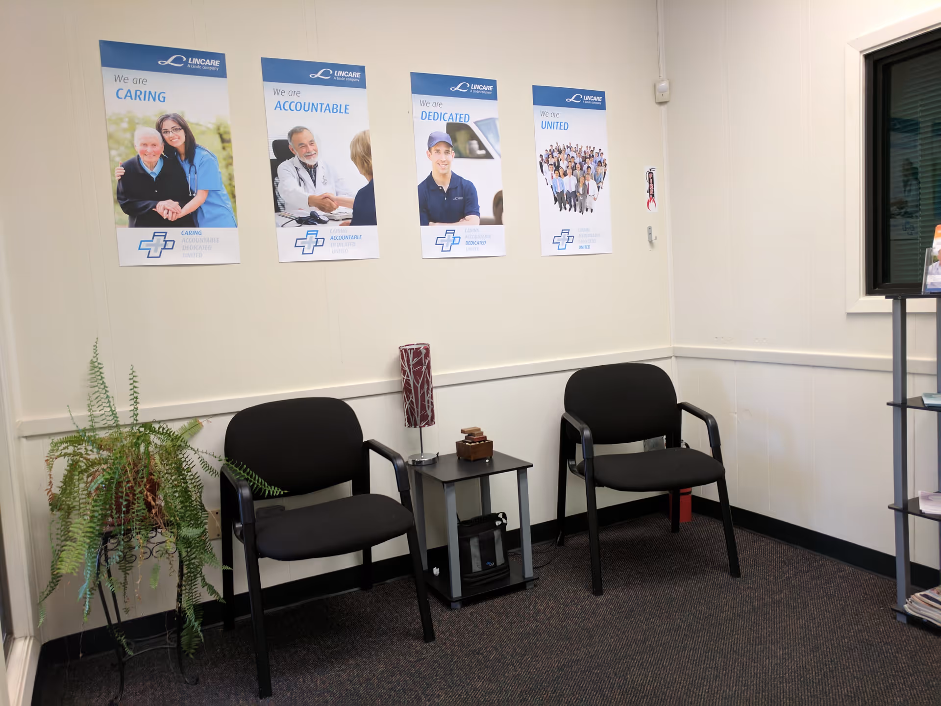 Indoor waiting area with two black chairs, a small table, fern, and posters.