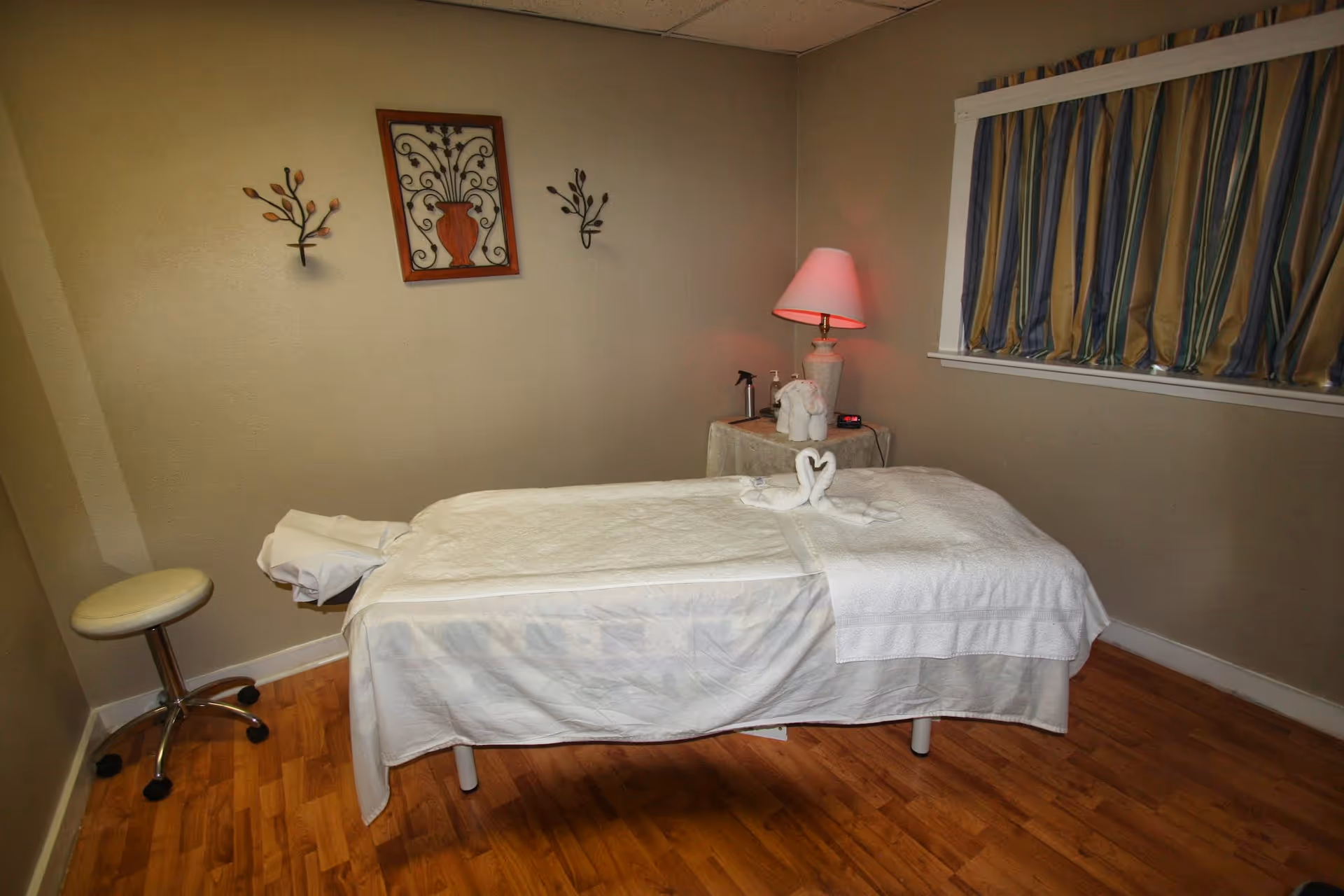 Treatment room with a massage bed, pink lamp, window curtains, and swan towels.