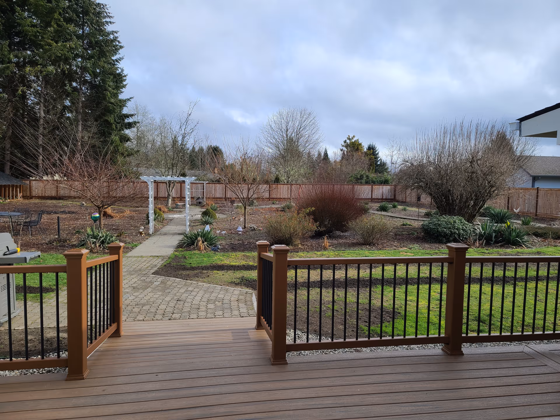 Wooden deck overlooks a landscaped backyard with a stone path, pergola, trees, and garden beds.