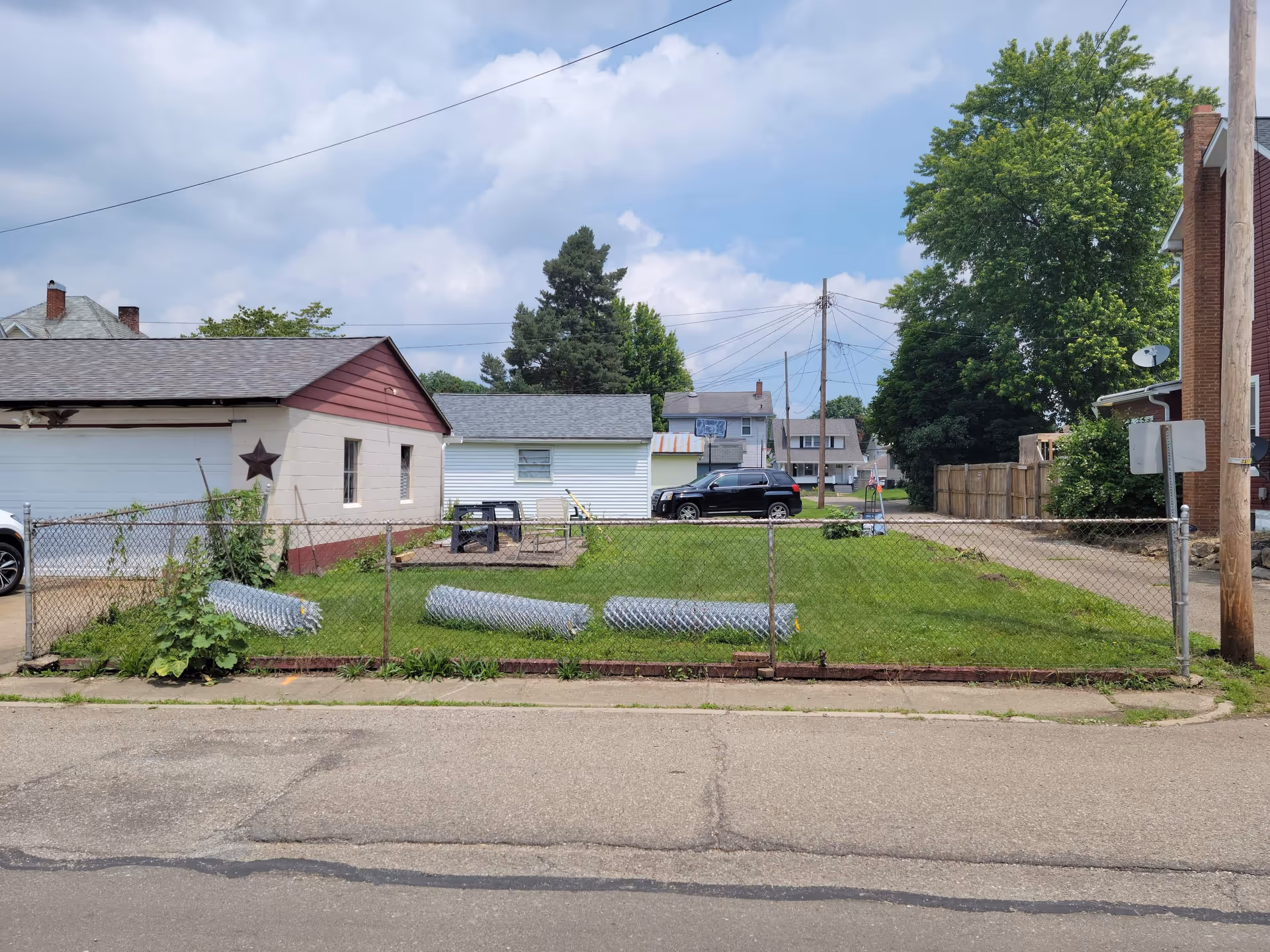 Exterior view of a fenced grassy lot with small buildings along a street.