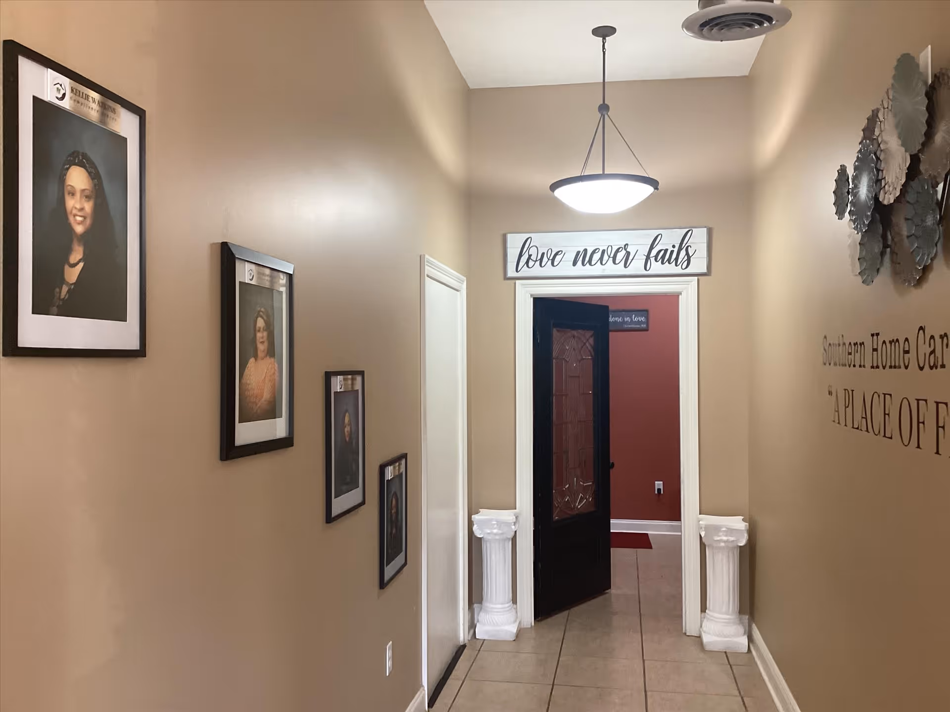 A hallway with framed portraits on the left and a doorway with 'love never fails' sign.