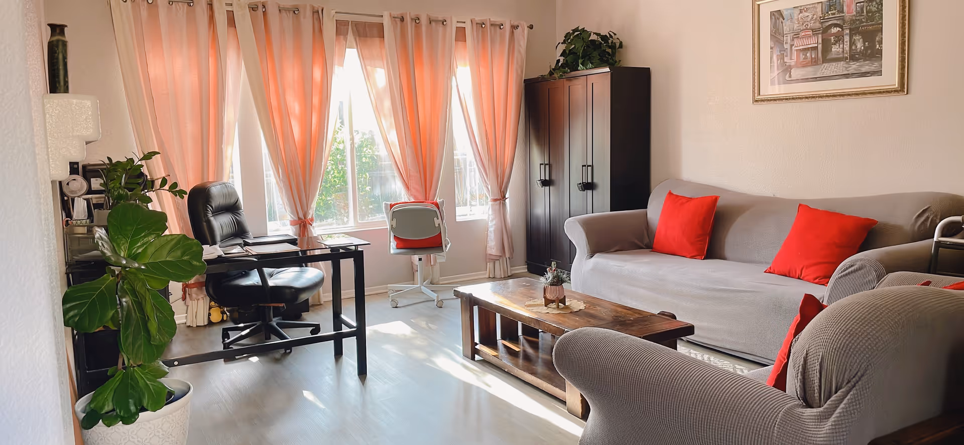 Cozy living room with gray sofa, red cushions, coffee table, desk, and pink-curtained windows.
