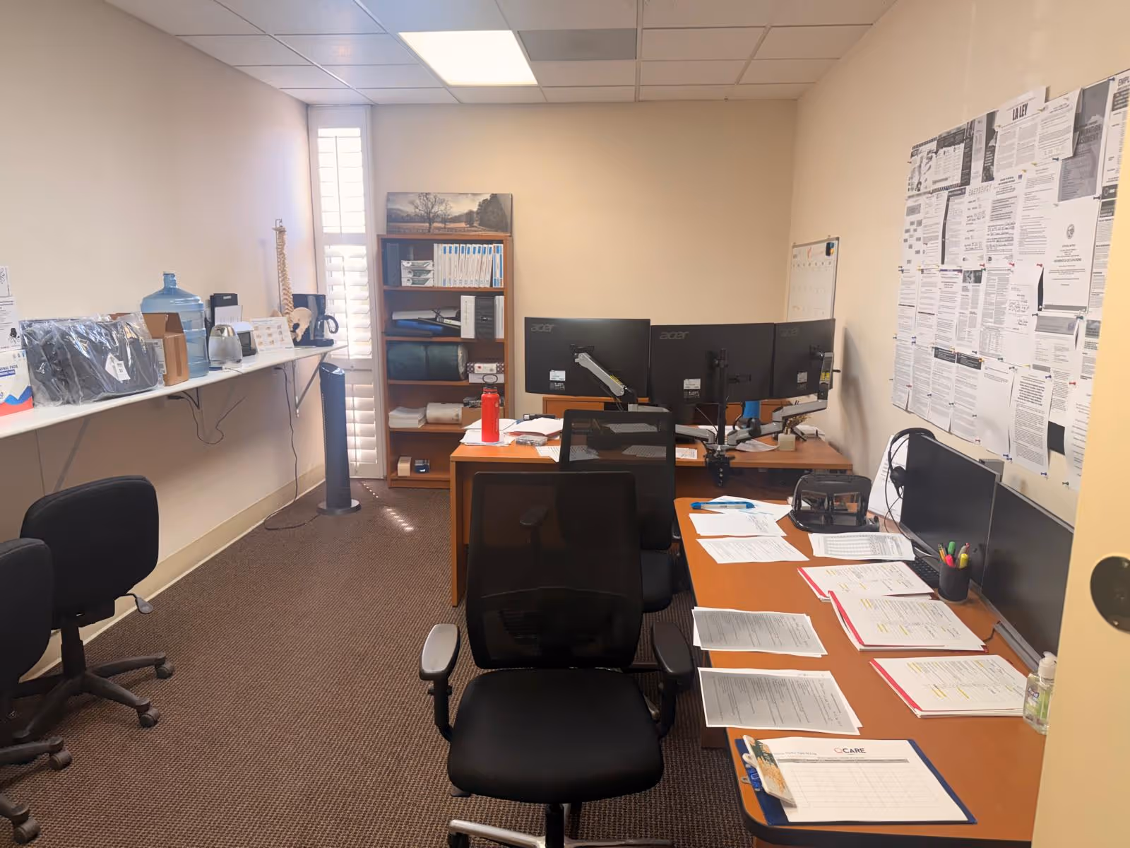 Office workspace with desks, dual monitors, paperwork, and chairs in a small facility.