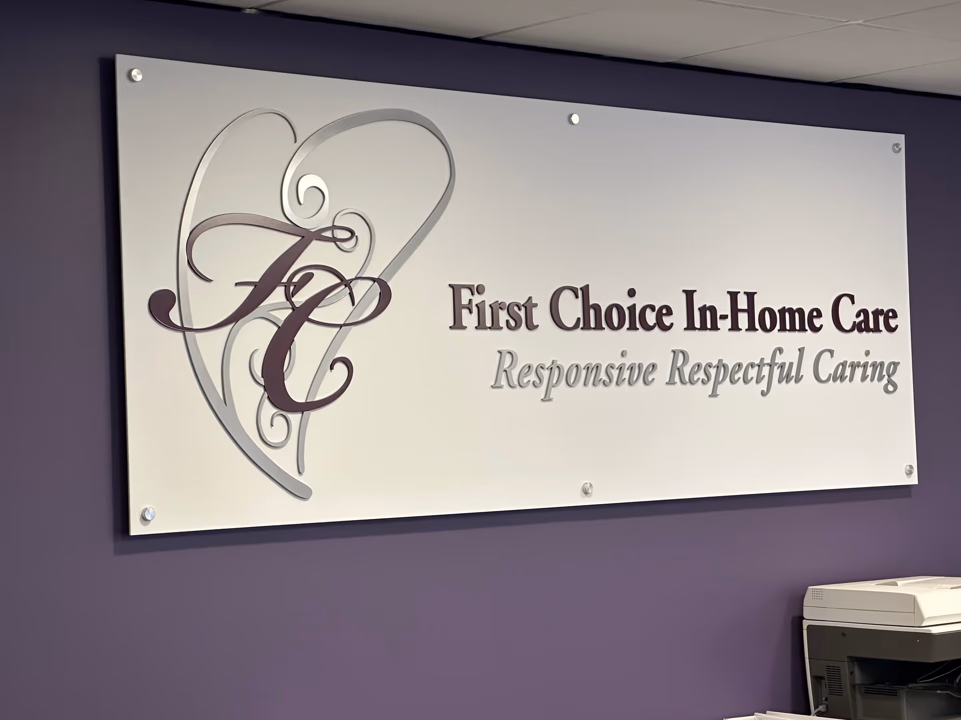 Metal sign with 'First Choice In-Home Care' logo mounted on purple wall beside a printer.