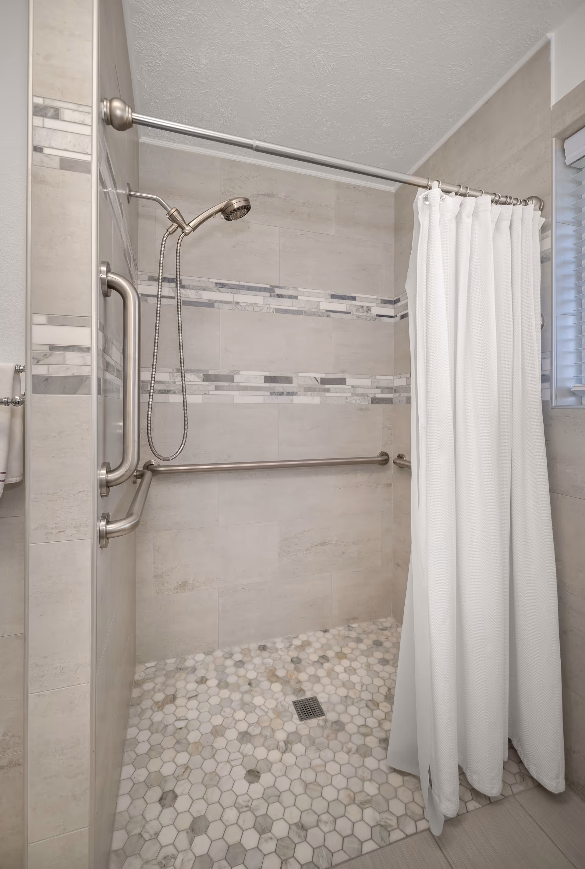 Accessible shower area in beige tiled bathroom with grab bars and a white curtain.