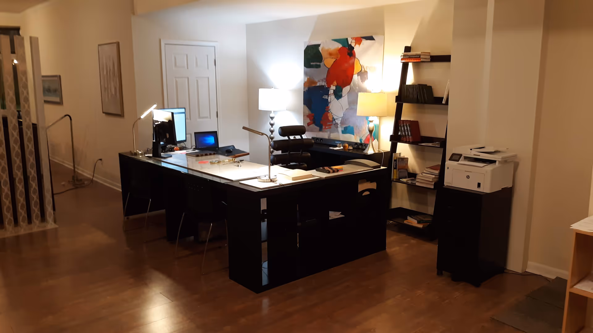 Interior office space with a large L-shaped desk, computer, lamps, and art on the wall.